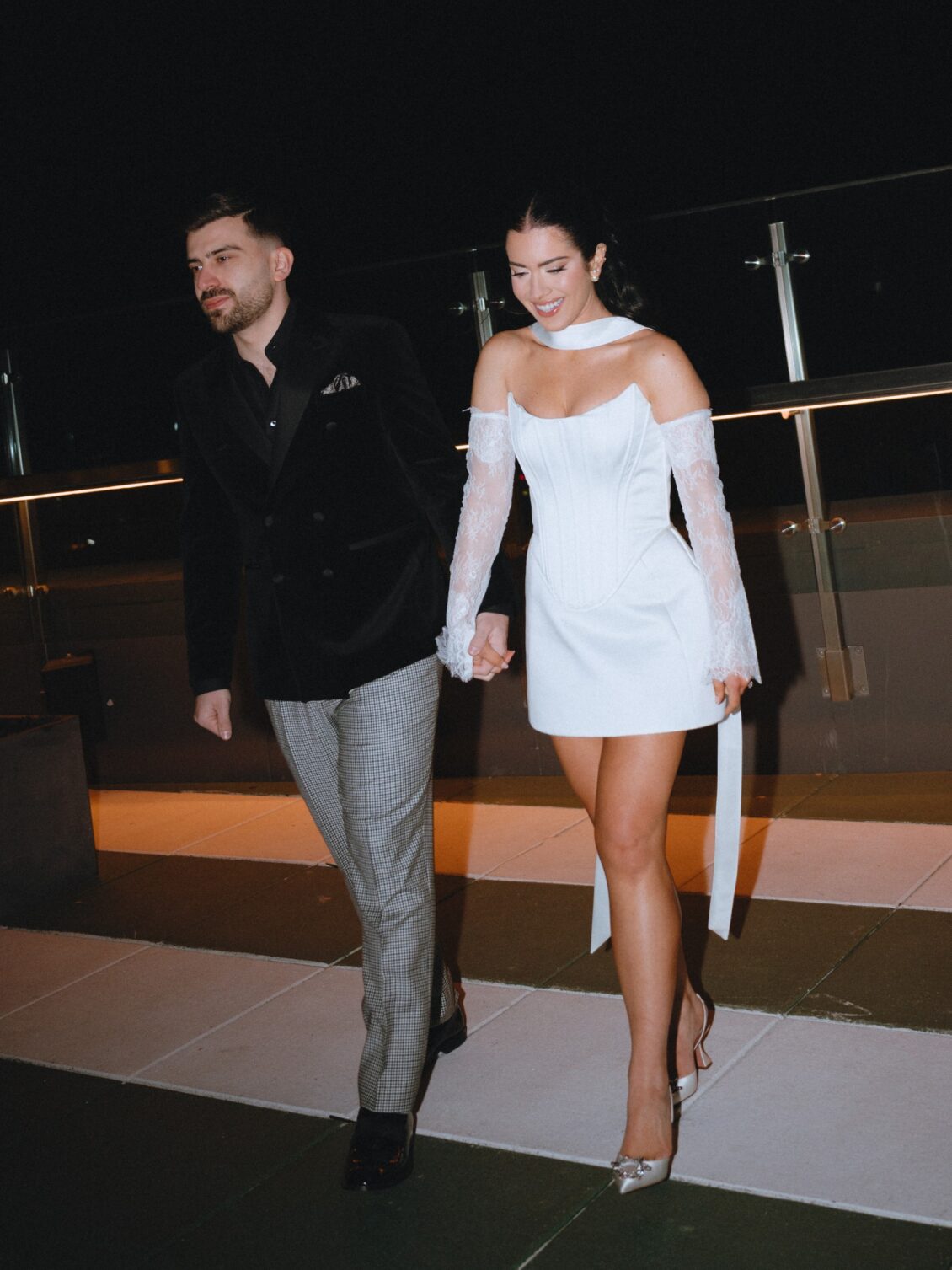 Valerie and Cosimo walk hand in hand across a tiled terrace at night in black-and-white cocktail attire.