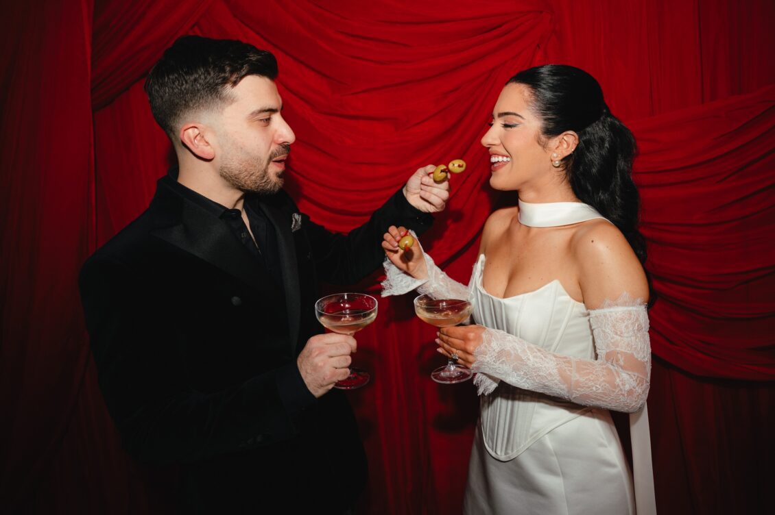 Valerie and Cosimo smile at each other while offering green cocktail olives across coupe glasses before red drapery.