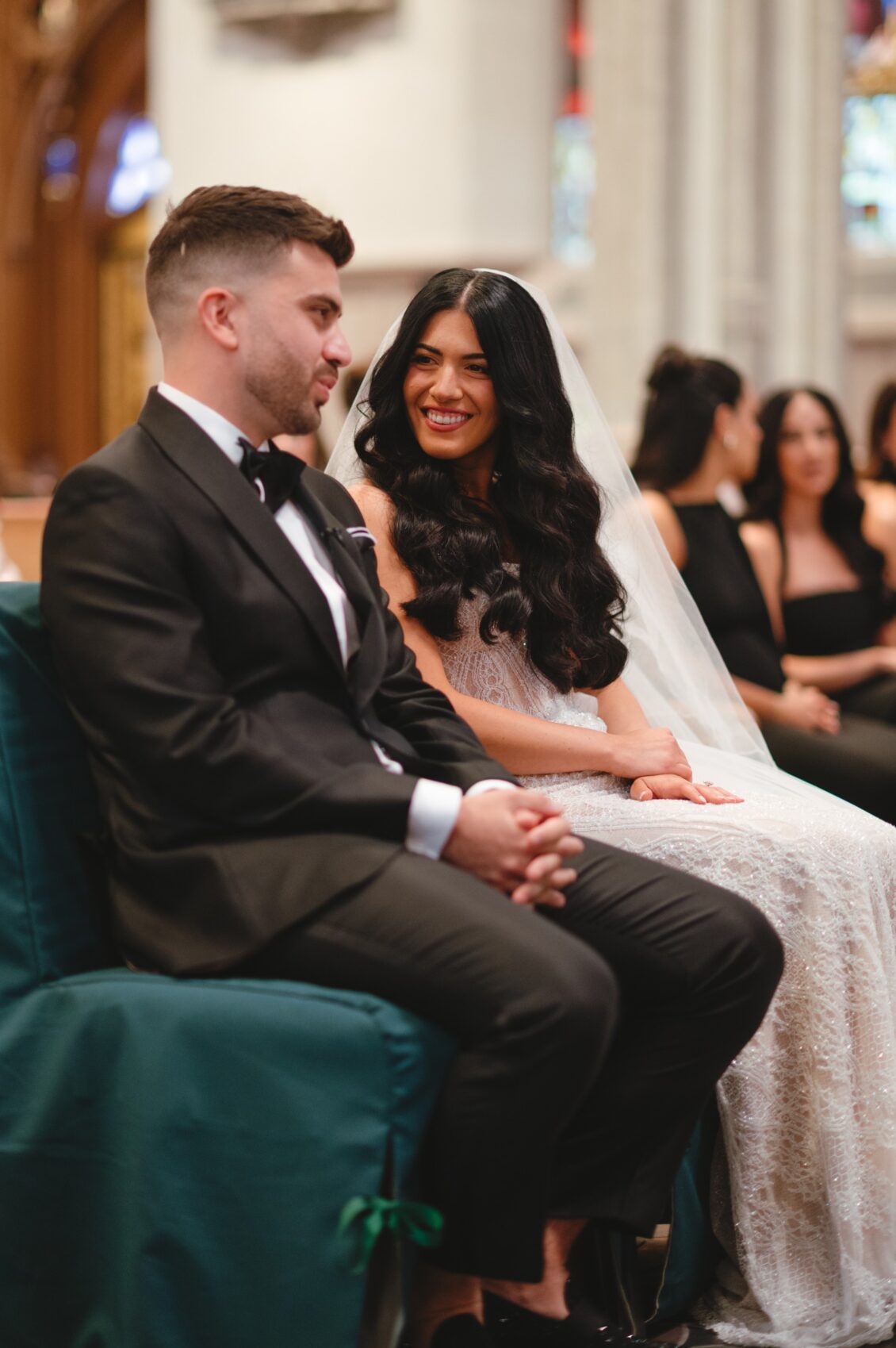 Valerie and Cosimo smile at each other while seated hand in hand on a dark green chair inside the cathedral.