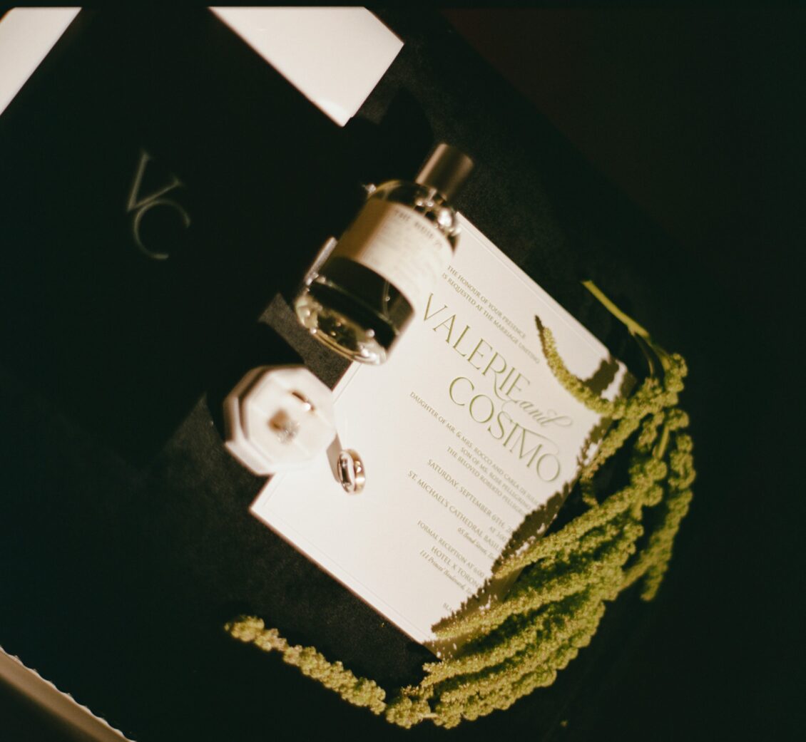A cream Valerie and Cosimo invitation lies with a clear perfume bottle, gold-green strands, and a black VC monogram piece.