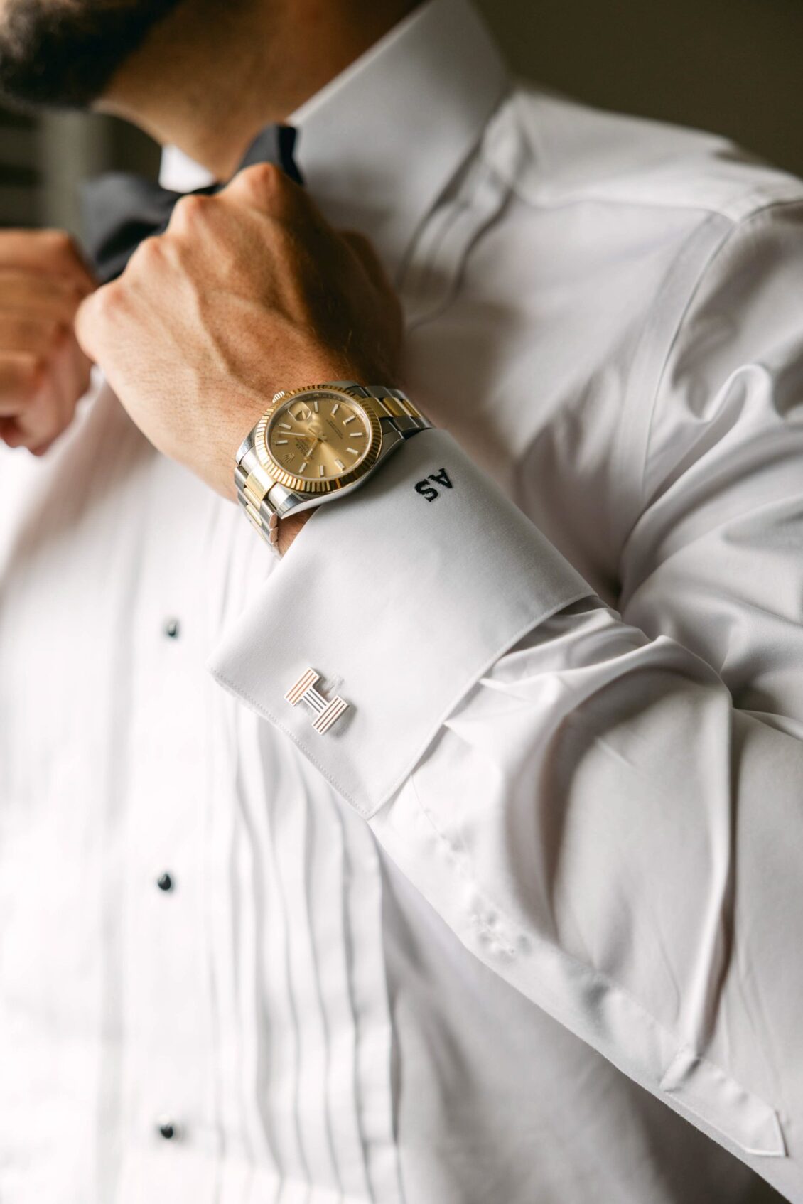 Alex fastening the monogrammed French cuff of his white tuxedo shirt, a two-tone watch on his wrist