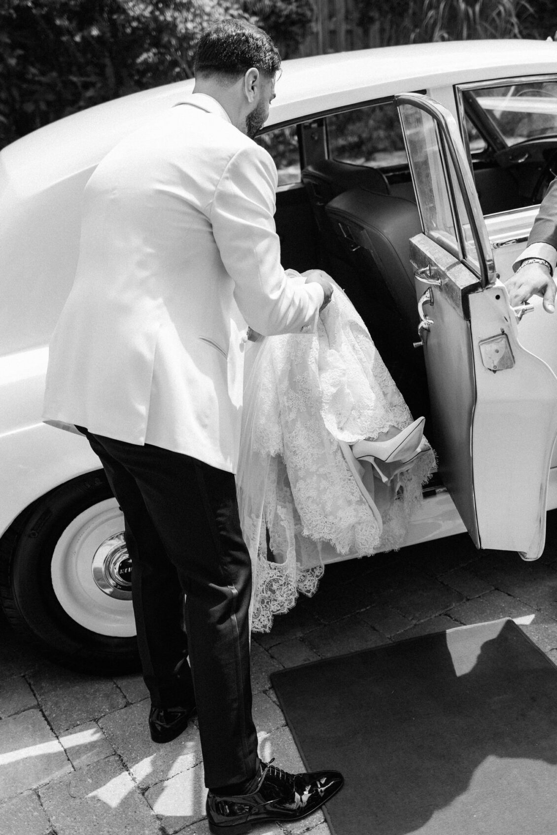 Black and white photo of Alex gathering Lara's lace gown and veil as she steps into a white vintage car