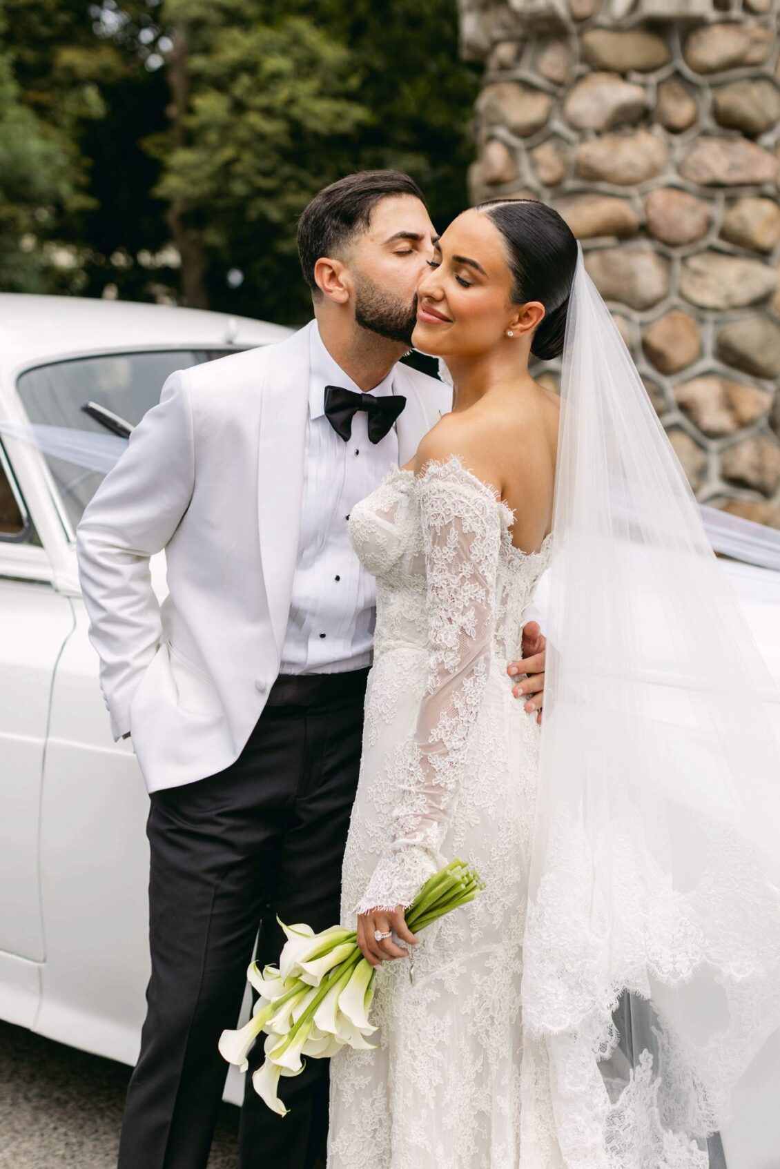 Alex kisses Lara's cheek beside a white vintage car and a stone wall