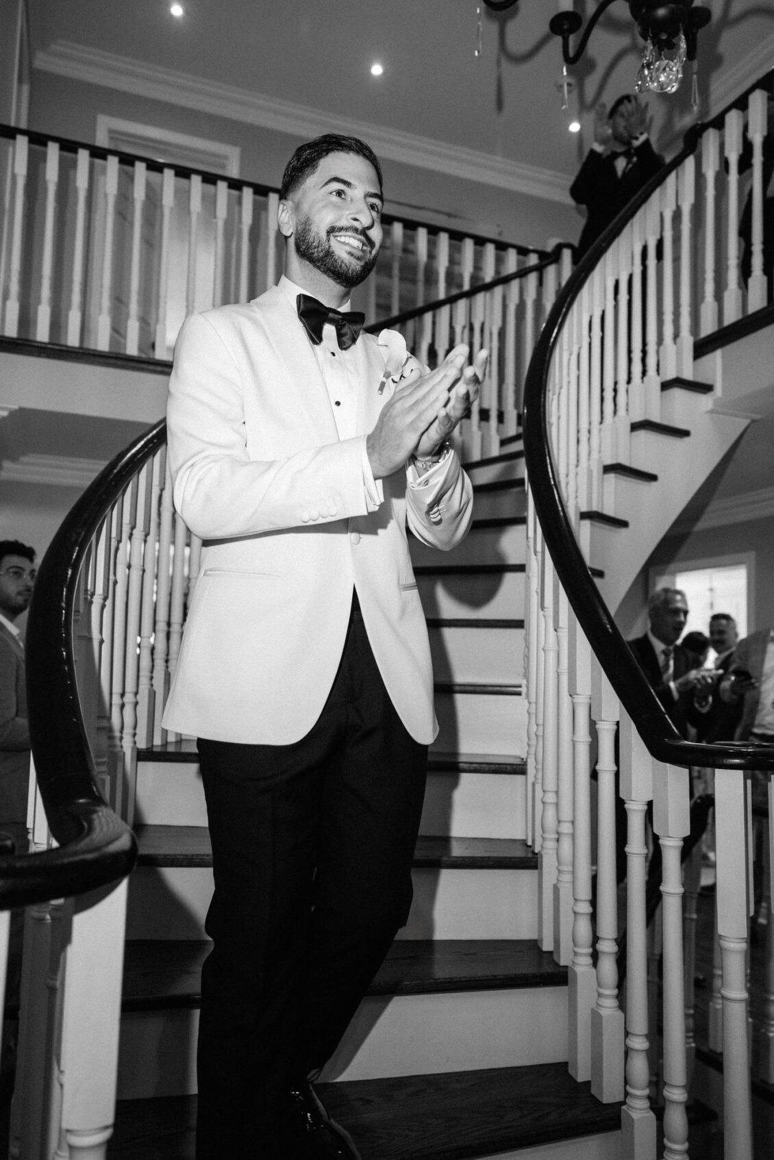 Black and white photo of Alex in a white tuxedo jacket and black bow tie clapping on a curved staircase