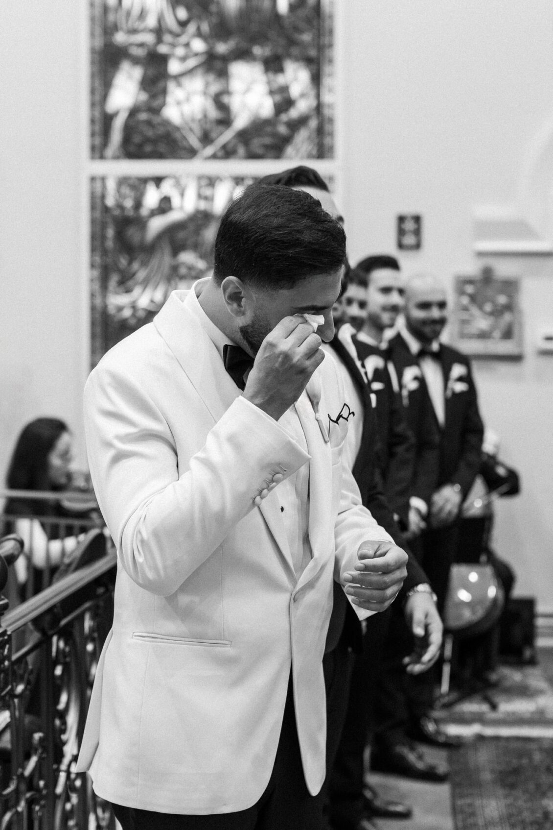 Black and white photo of Alex in a white tuxedo jacket wiping a tear with his groomsmen behind him