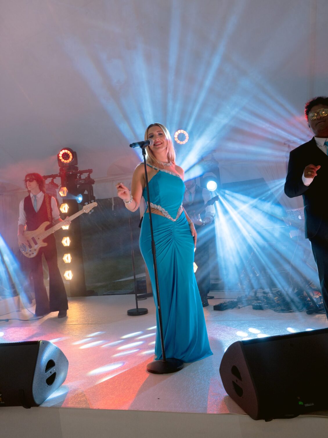 A singer in a teal gown performs beneath blue stage lights with the band behind her