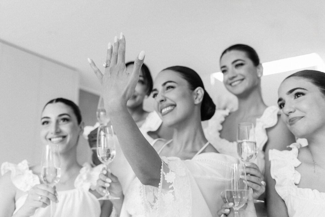 Black and white photo of Lara and her bridesmaids in white ruffled dresses holding champagne as Lara raises her ring hand