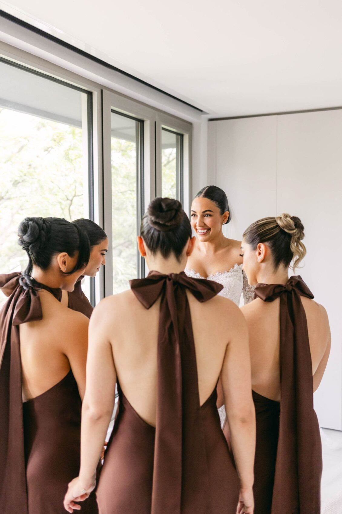 Bridesmaids seen from behind in chocolate-brown halter gowns tied with large bows as Lara smiles at them
