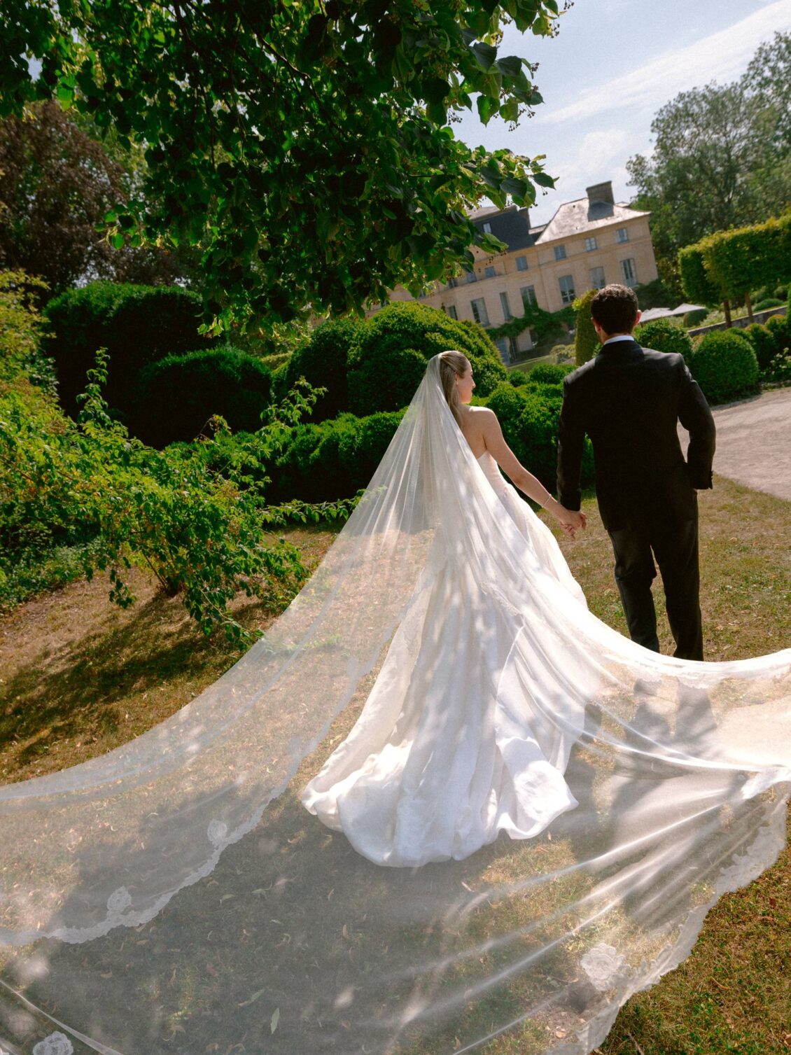 Lamya's long veil and train spread across the grass as she faces Sam, with the château behind them