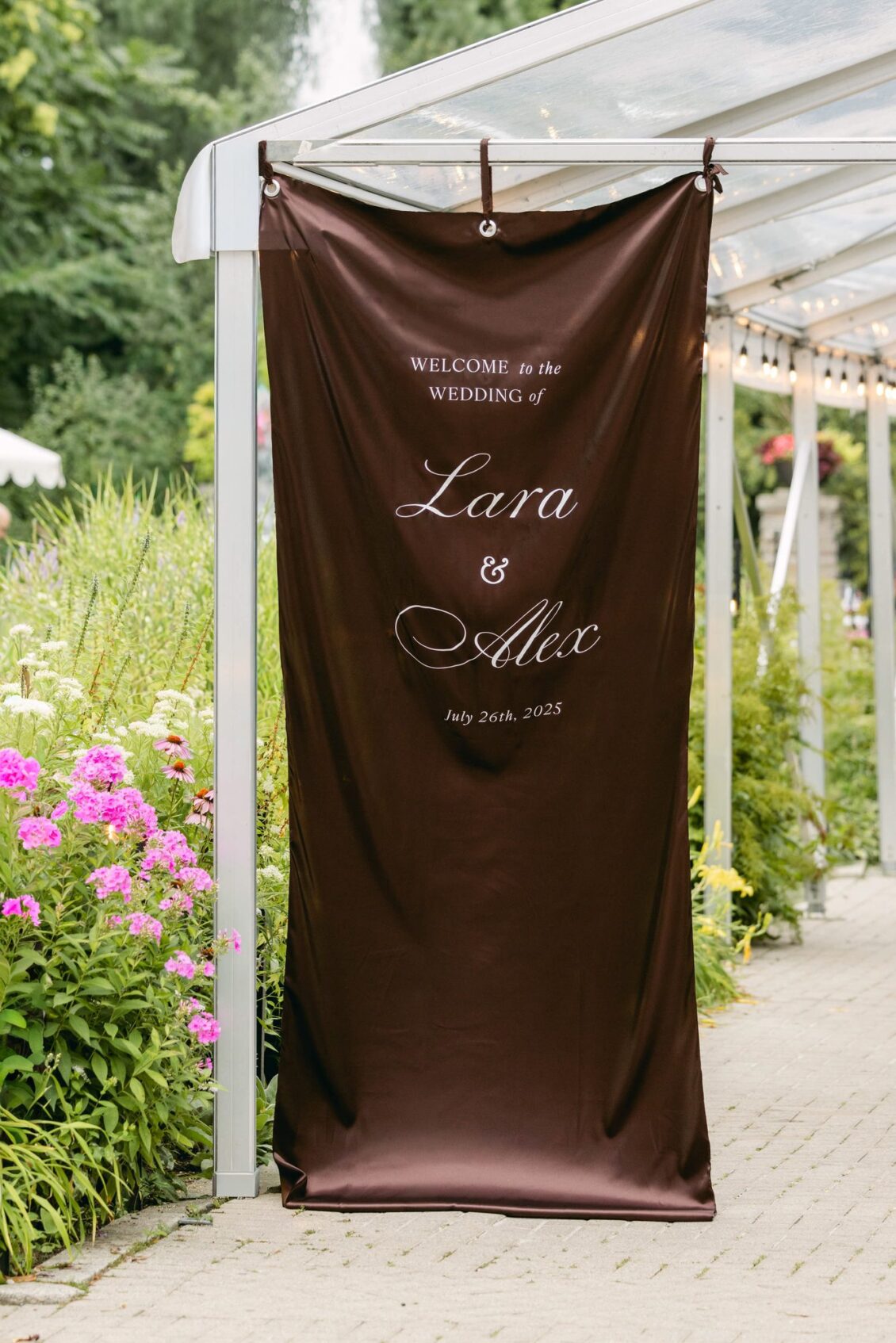 Chocolate-brown fabric banner welcoming guests to the wedding of Lara and Alex hanging in a garden pergola