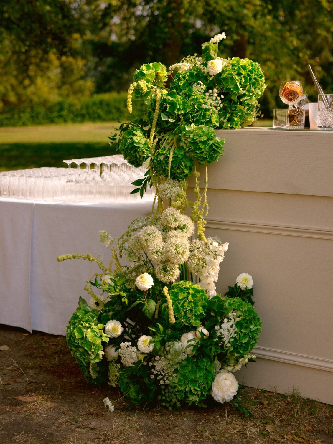 A cocktail bar dressed with a column of green and white flowers and rows of champagne coupes