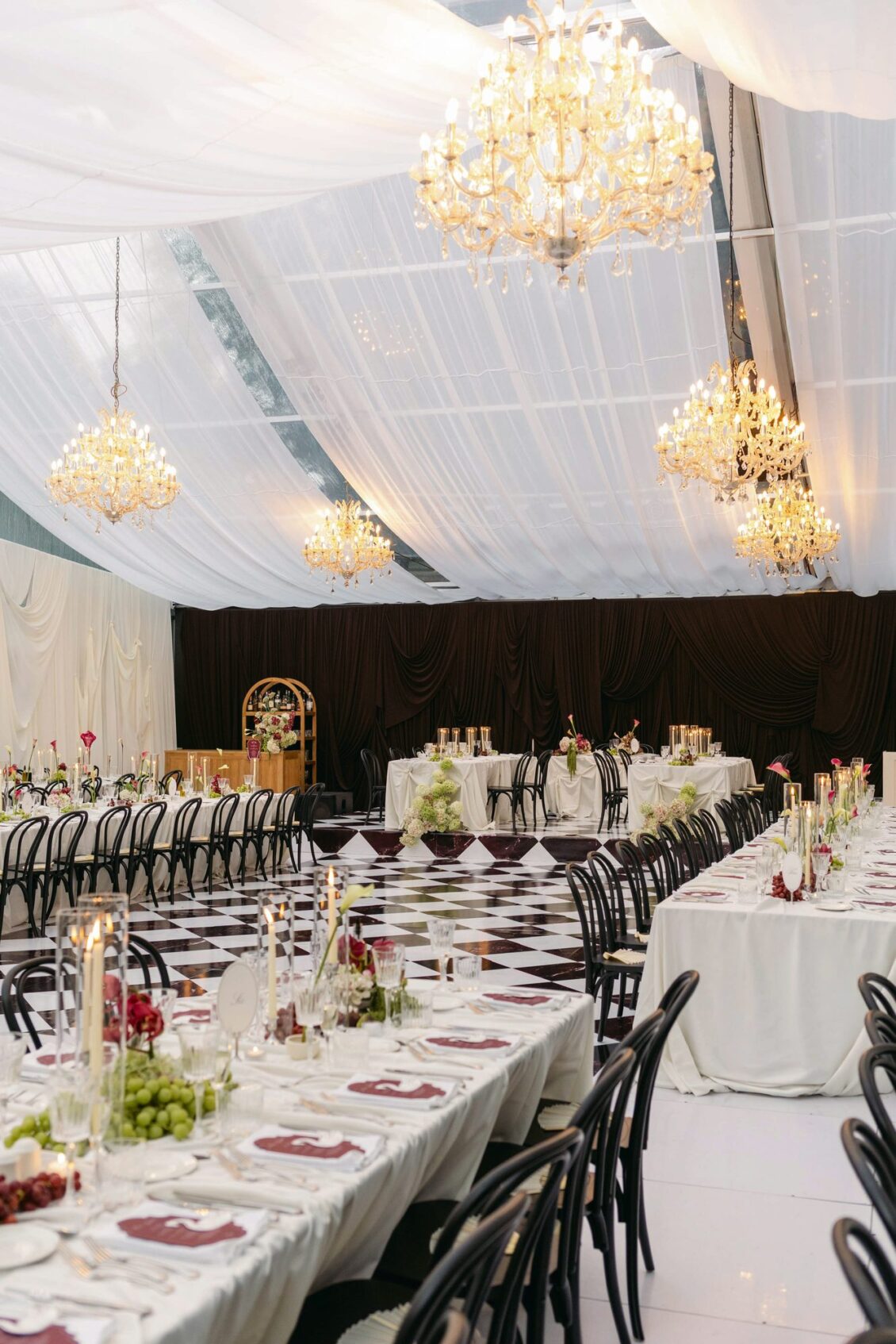 Wide view of the draped reception pavilion with crystal chandeliers, long tables and a checkered floor
