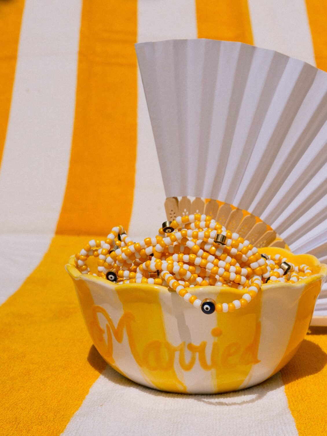 A hand-painted bowl reading Married filled with yellow-and-white evil-eye bracelets beside a paper fan