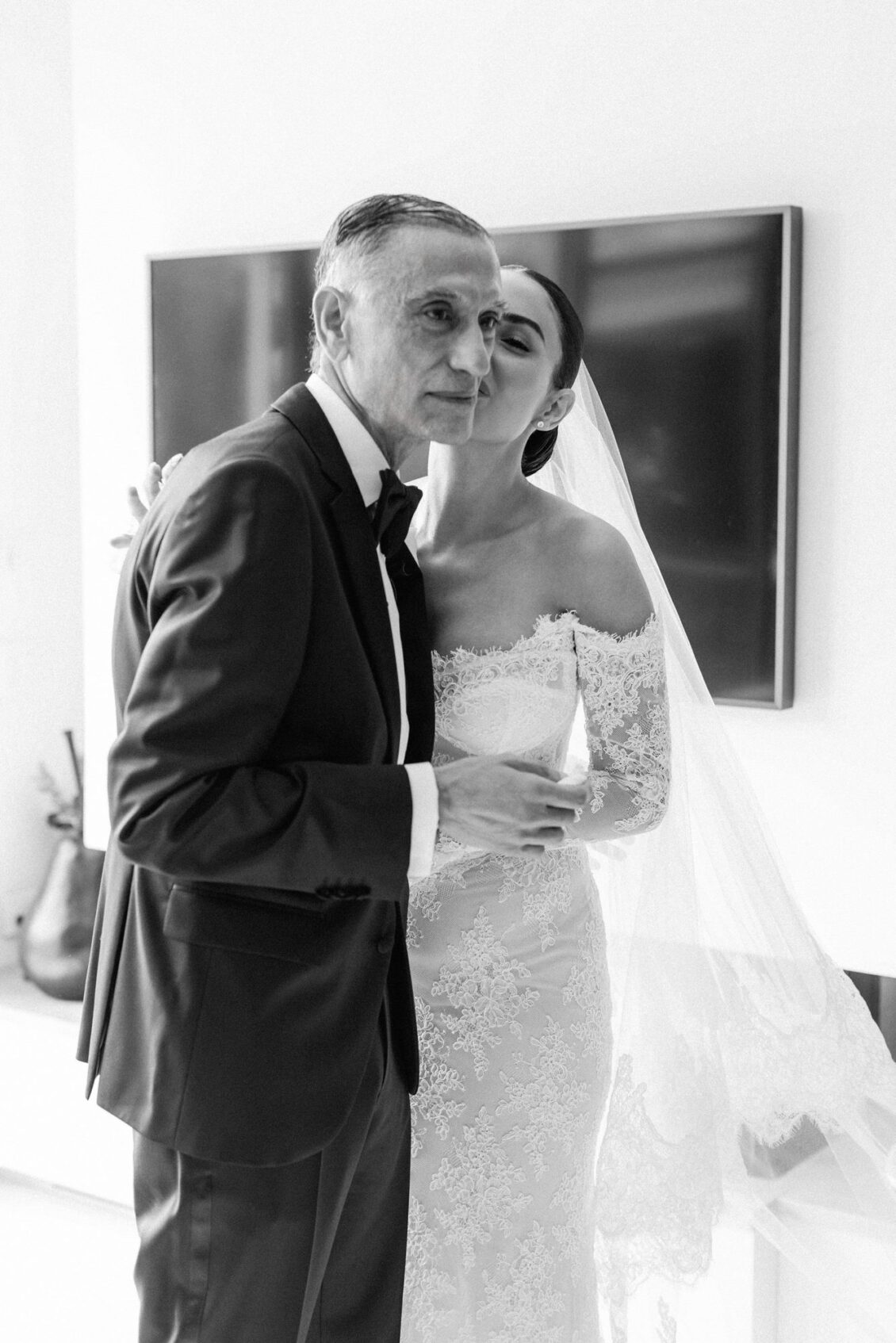 Lara's father kisses her cheek as she smiles in her lace gown and veil