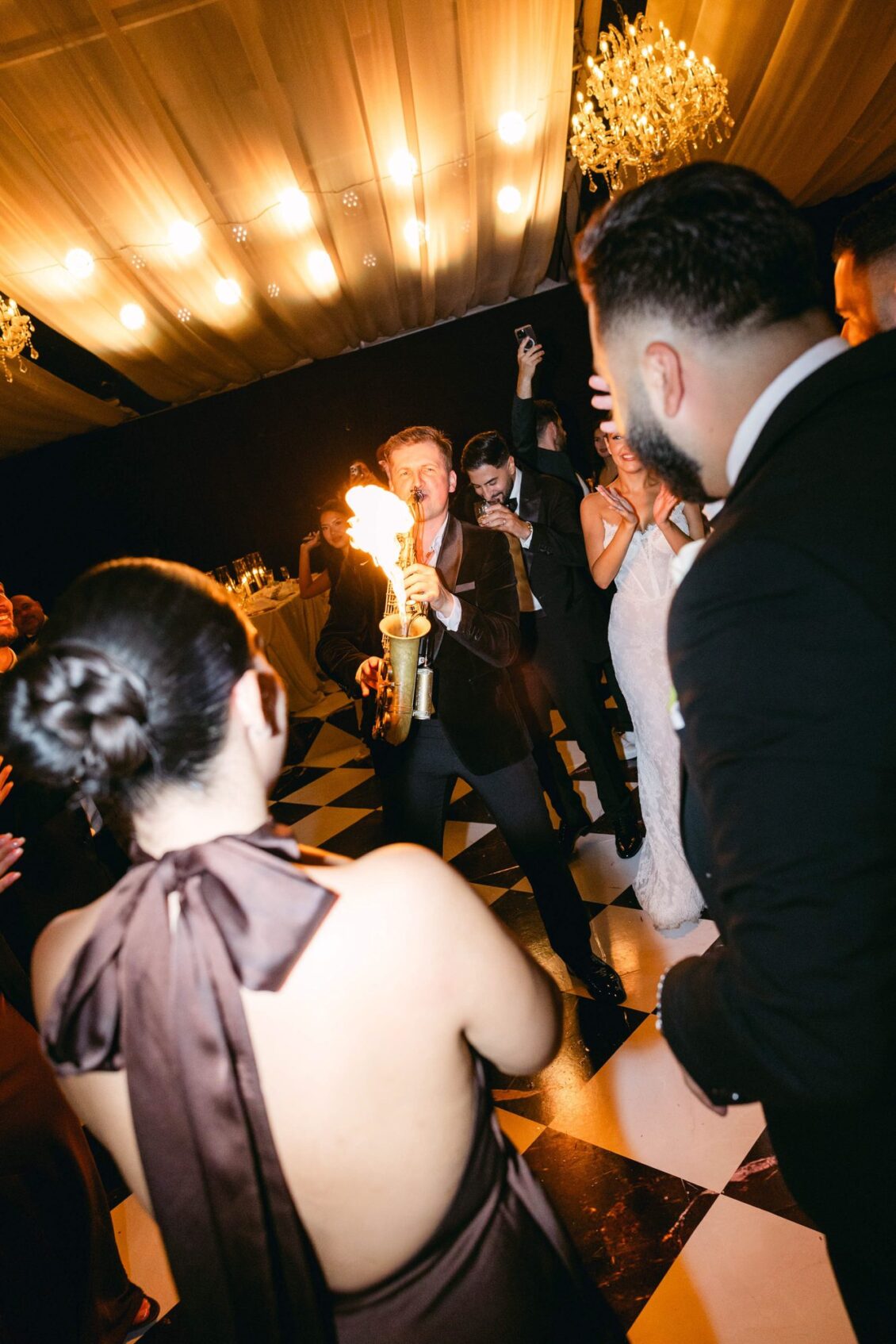 A saxophonist plays a flaming saxophone on the checkered dance floor as Lara and guests cheer behind him