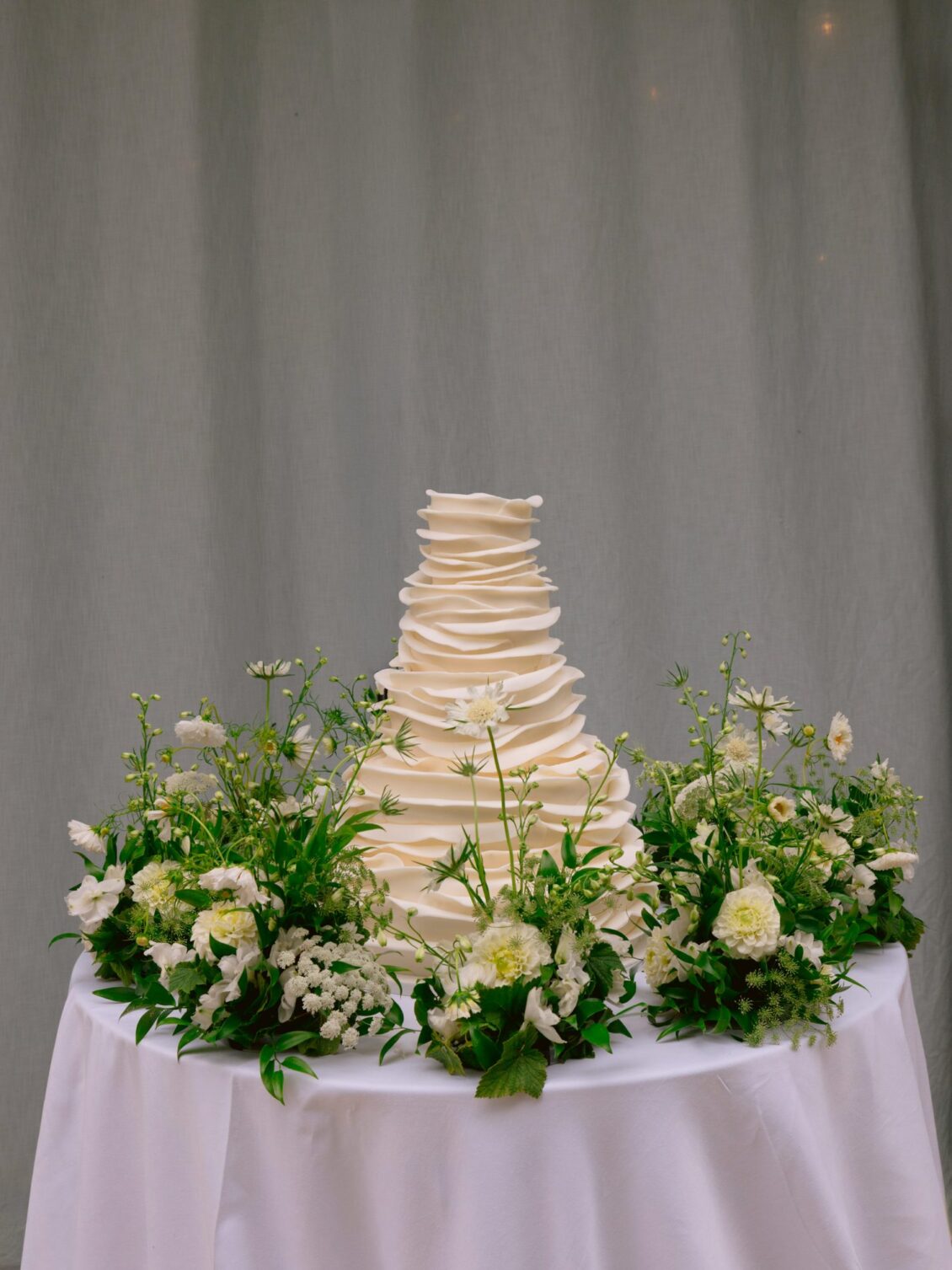 A tiered white ruffled wedding cake ringed with white garden flowers on a draped table