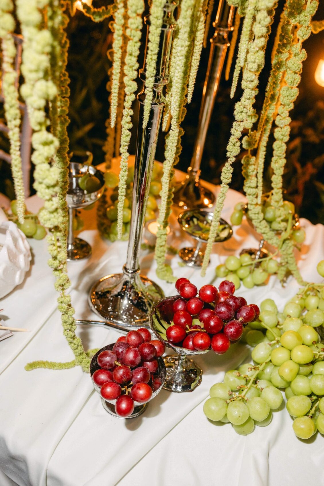 Red and green grapes in silver dishes on a white table beneath hanging green amaranthus
