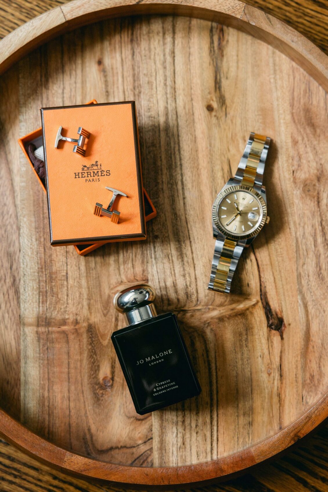 Cufflinks in an orange box, a two-tone watch and a bottle of cologne on a round wooden tray