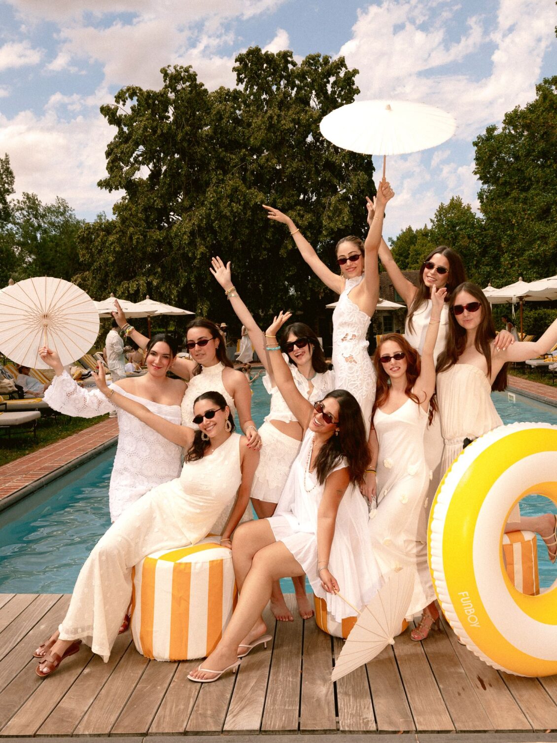 A group of women dressed in white pose with white parasols on the deck beside the pool