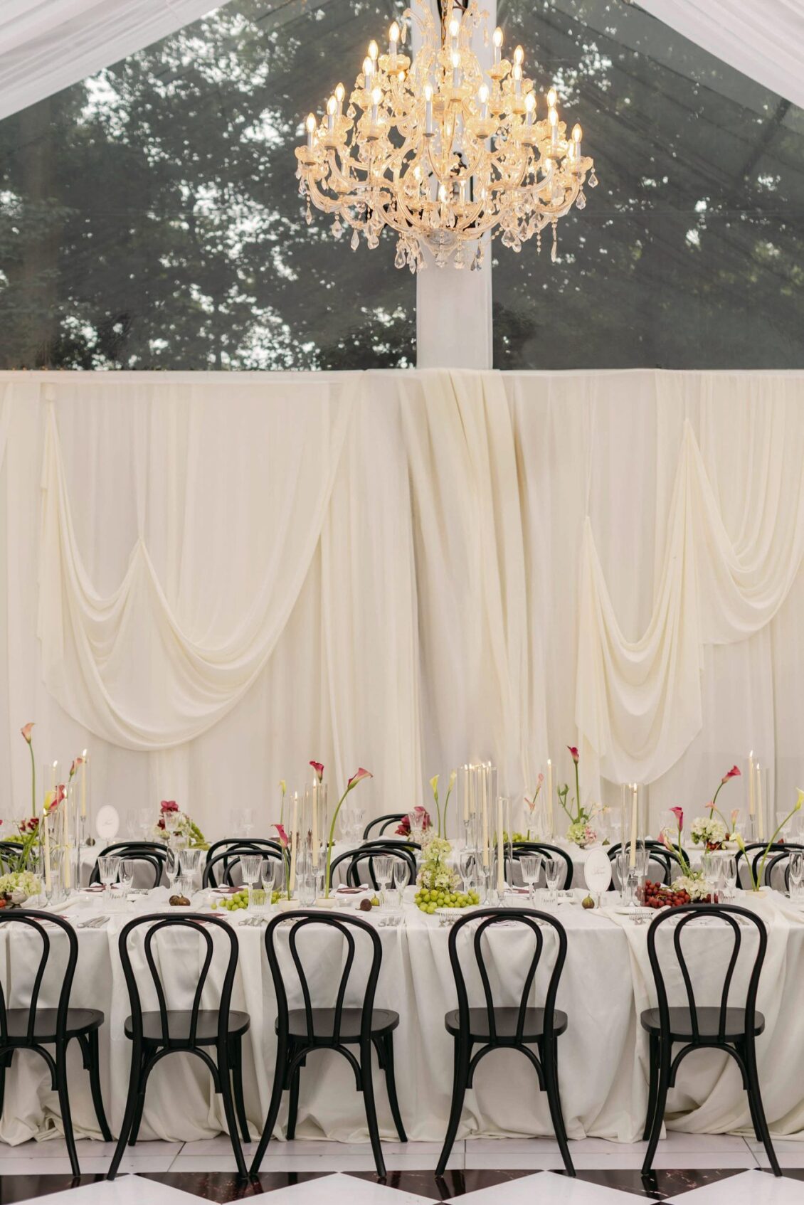 Long table with black bentwood chairs beneath swagged cream draping and a crystal chandelier