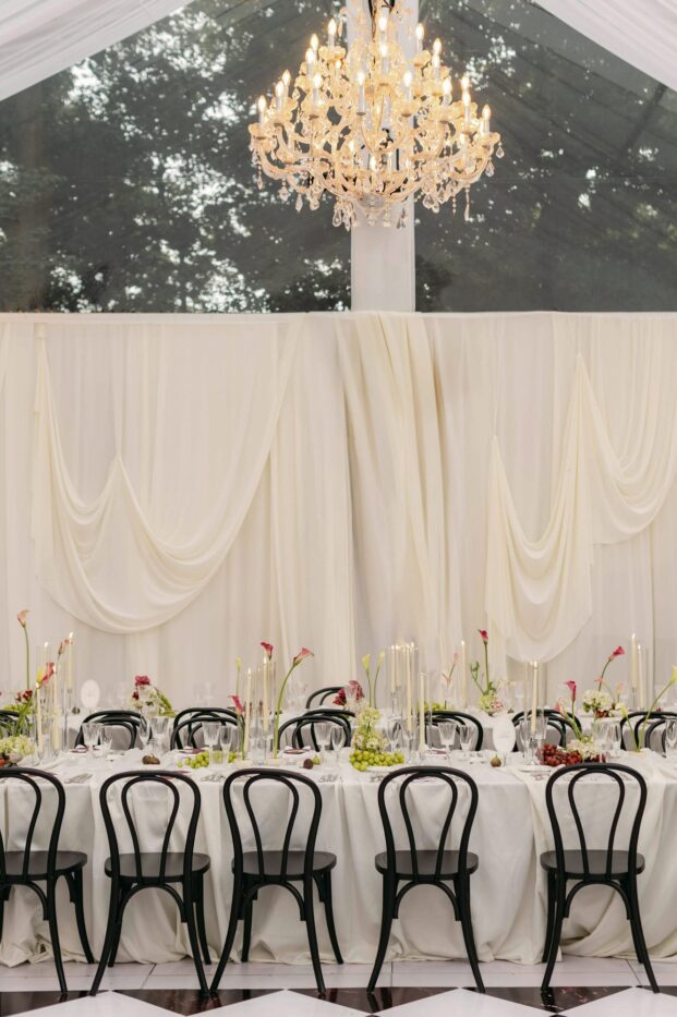 Long table with black bentwood chairs beneath swagged cream draping and a crystal chandelier