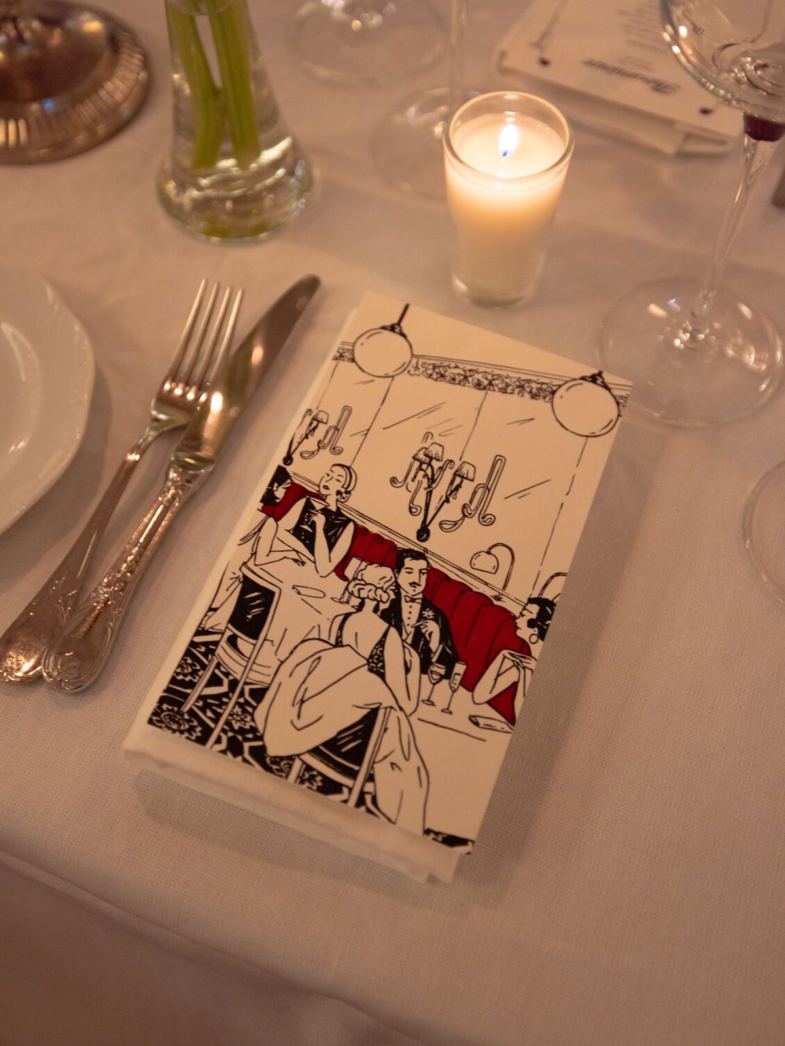 An illustrated black, white and red bistrot menu on a white tablecloth beside silver cutlery and a votive candle