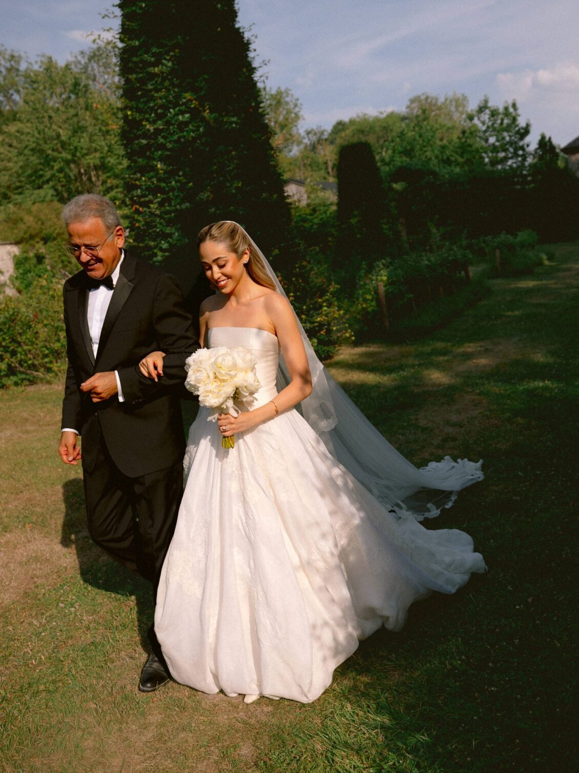 Lamya in a strapless gown and veil holds a white bouquet as she is escorted across the garden