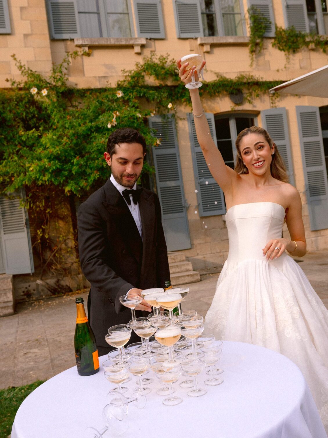 Lamya raises a coupe beside a champagne tower as Sam looks on in front of the château