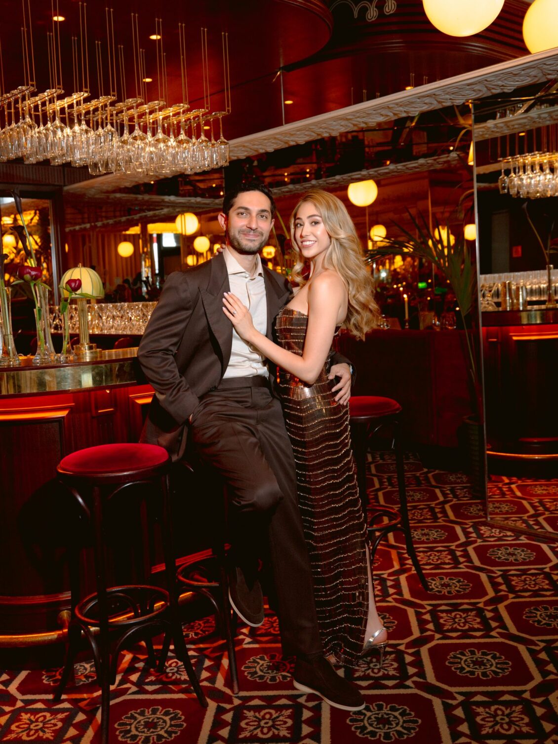 Lamya in a sparkling burgundy sequin gown stands with Sam in a tuxedo inside the red Thoumieux bistrot in Paris