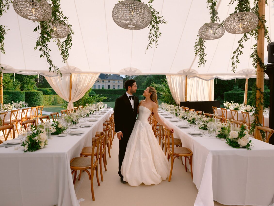 Lamya in a strapless gown and Sam in a tuxedo embrace between long white tables in a tent hung with greenery at Domaine de Primard