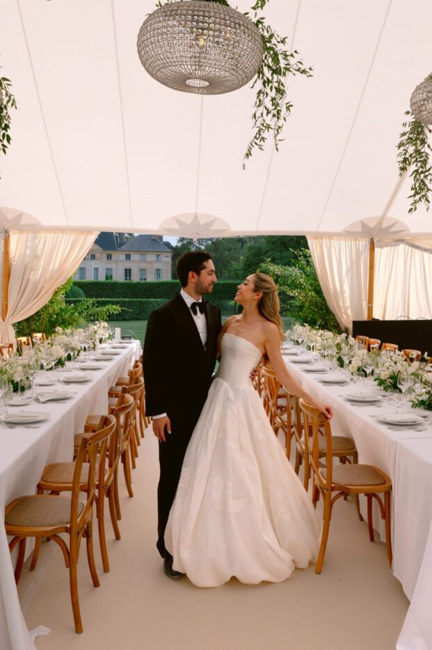 Lamya in a strapless gown and Sam in a tuxedo embrace between long white tables in a tent hung with greenery at Domaine de Primard
