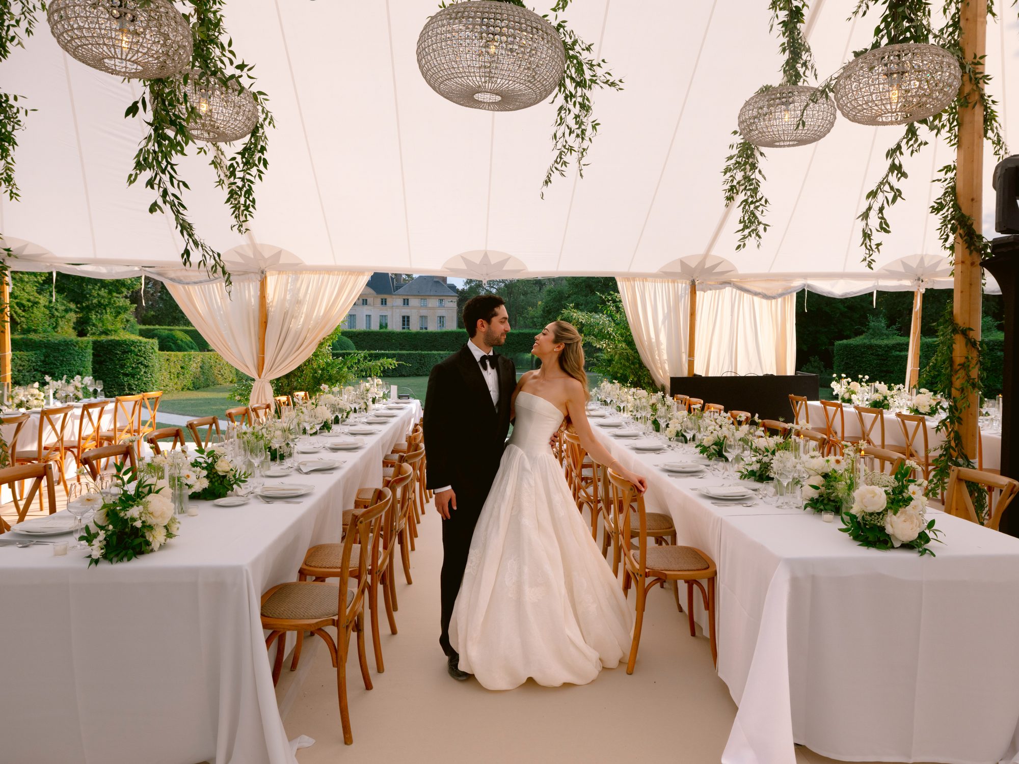 Lamya in a strapless gown and Sam in a tuxedo embrace between long white tables in a tent hung with greenery at Domaine de Primard