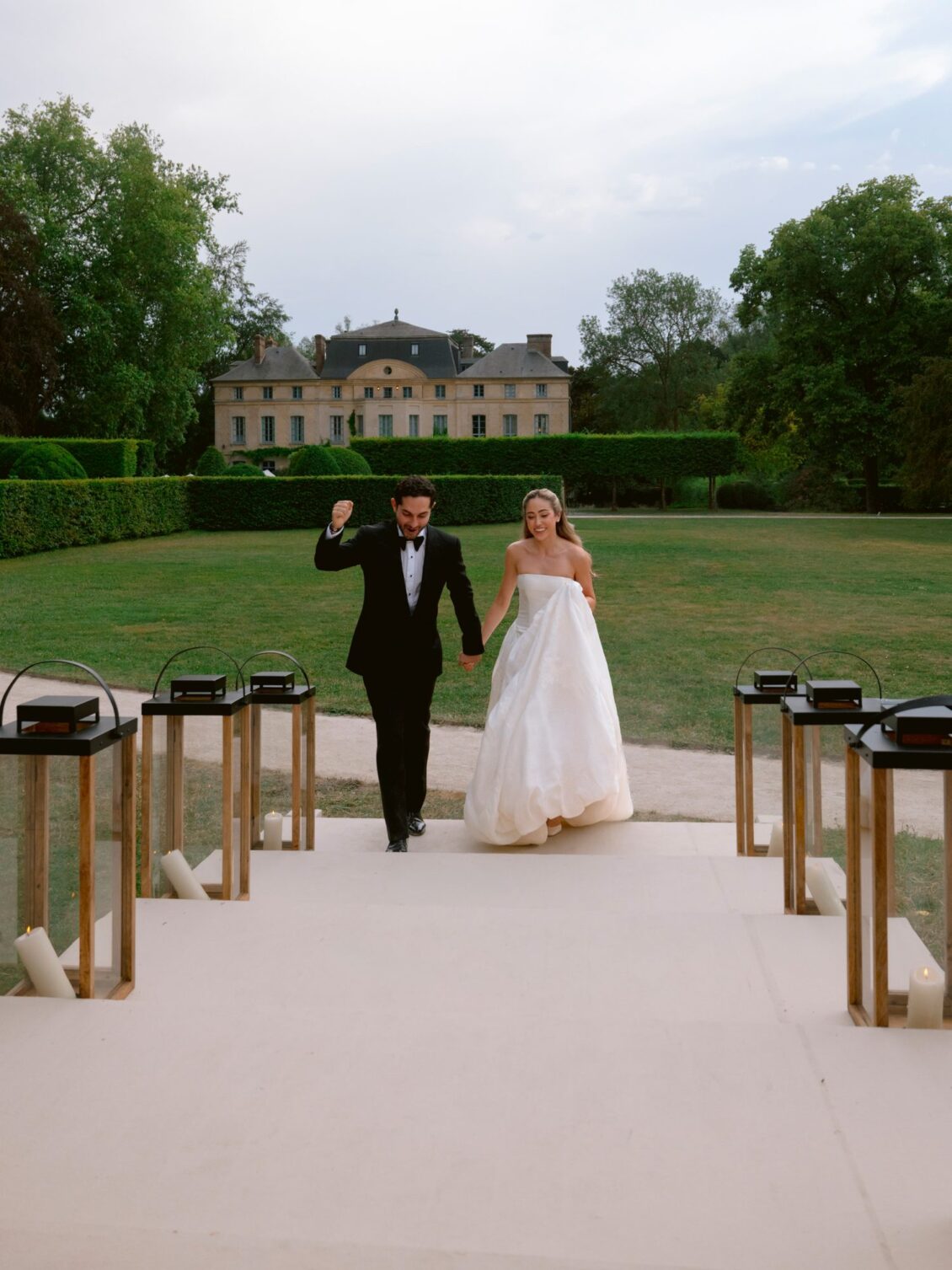 Lamya and Sam raise their arms as they walk the lantern-lined path to the tent, the château behind them