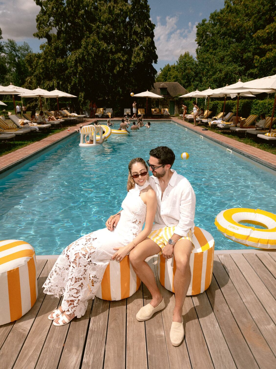 Lamya in a white lace dress and Sam in white sit together on striped poufs, the pool behind them