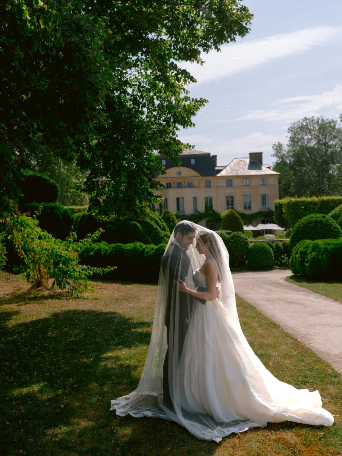 Lamya and Sam close together beneath her sheer veil on the lawn, with the château in the background