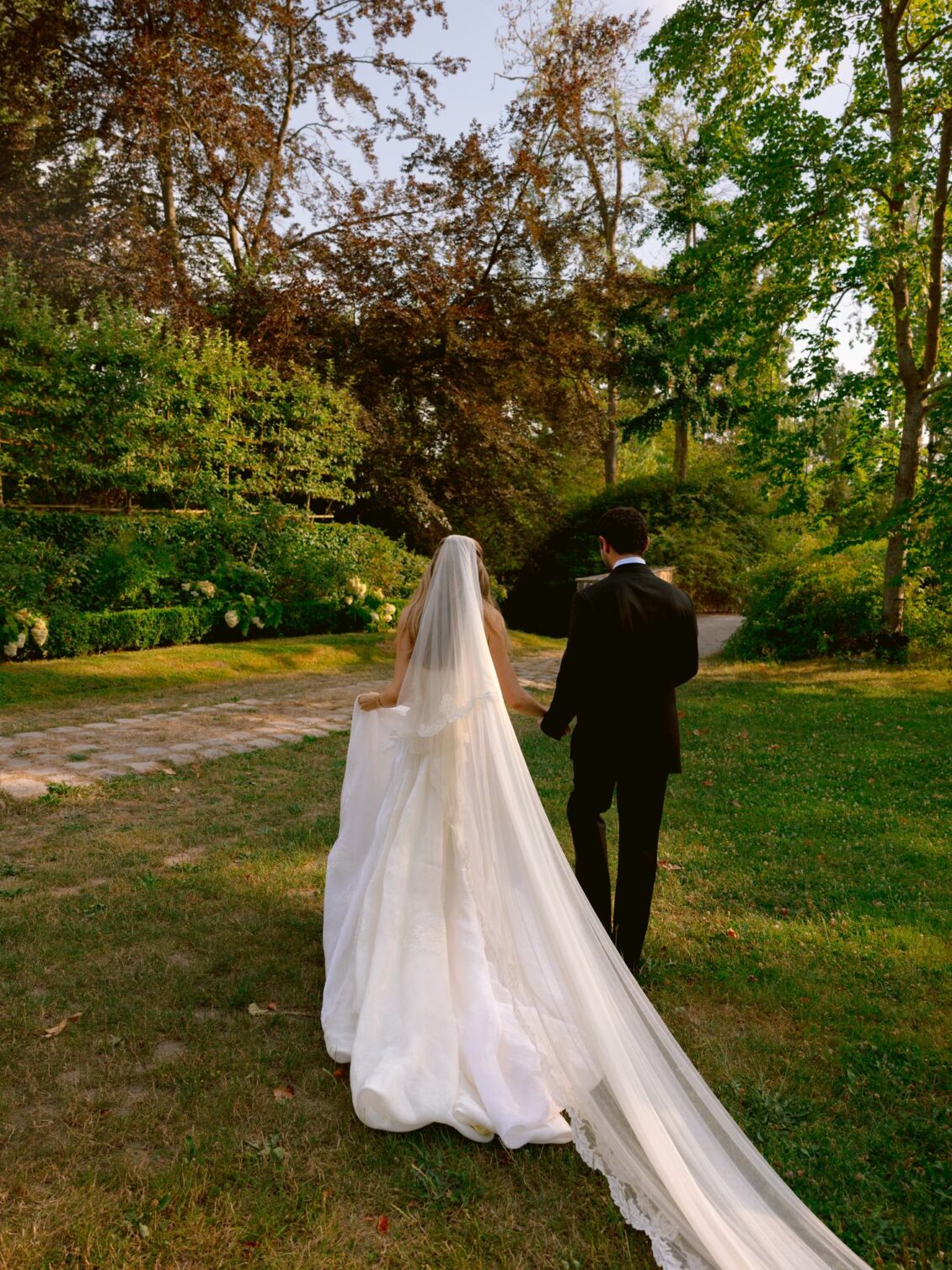 Lamya and Sam walk away across the lawn with her long veil trailing behind
