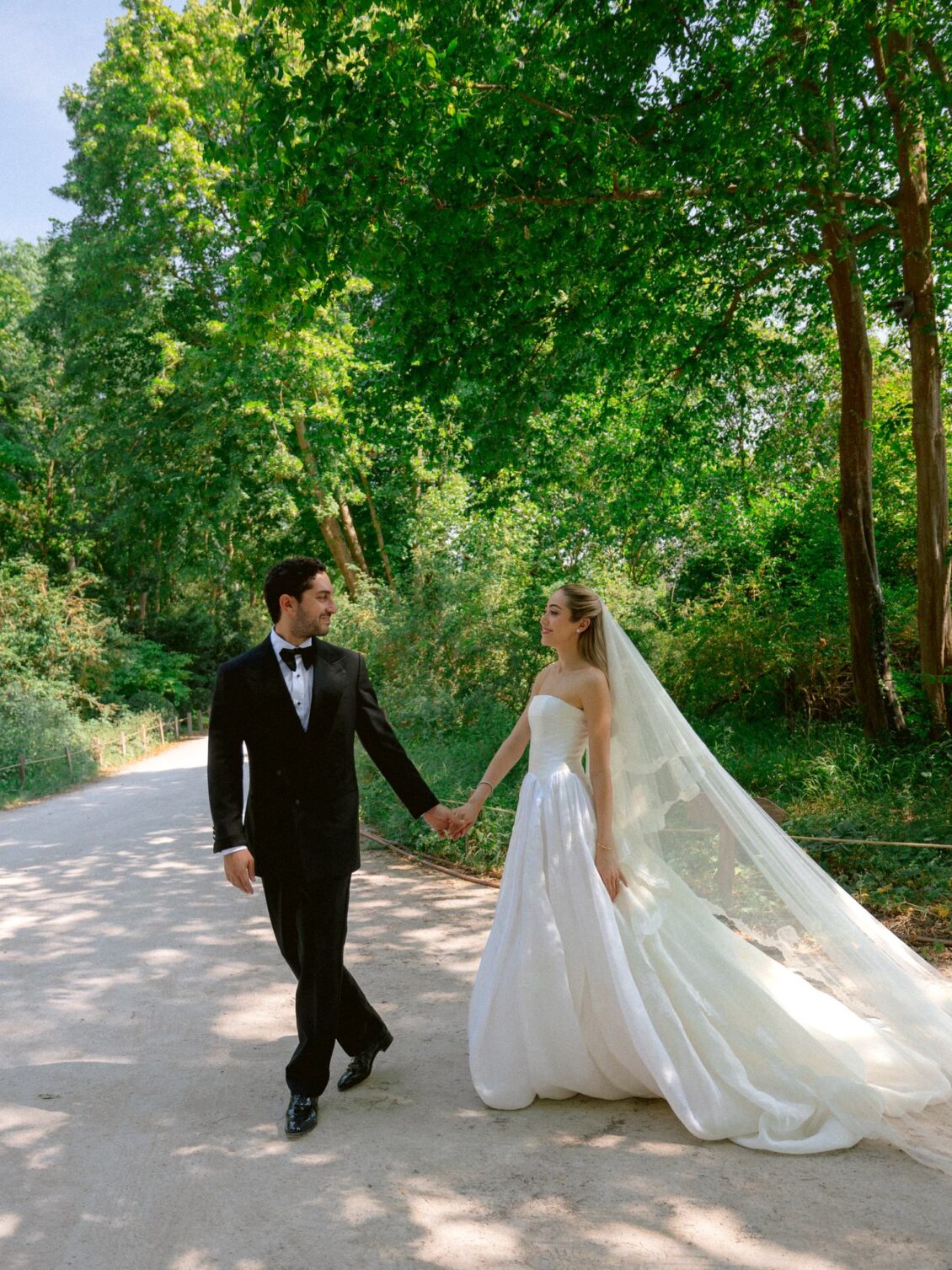 Lamya in a strapless gown and cathedral veil walks hand in hand with Sam down a sunlit tree-lined path