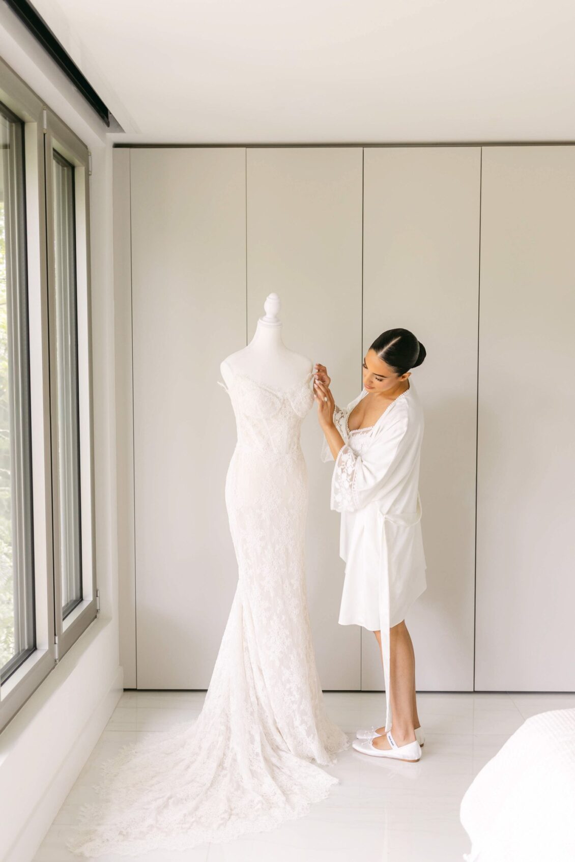 Lara in a white robe touching her lace wedding gown on a dress form