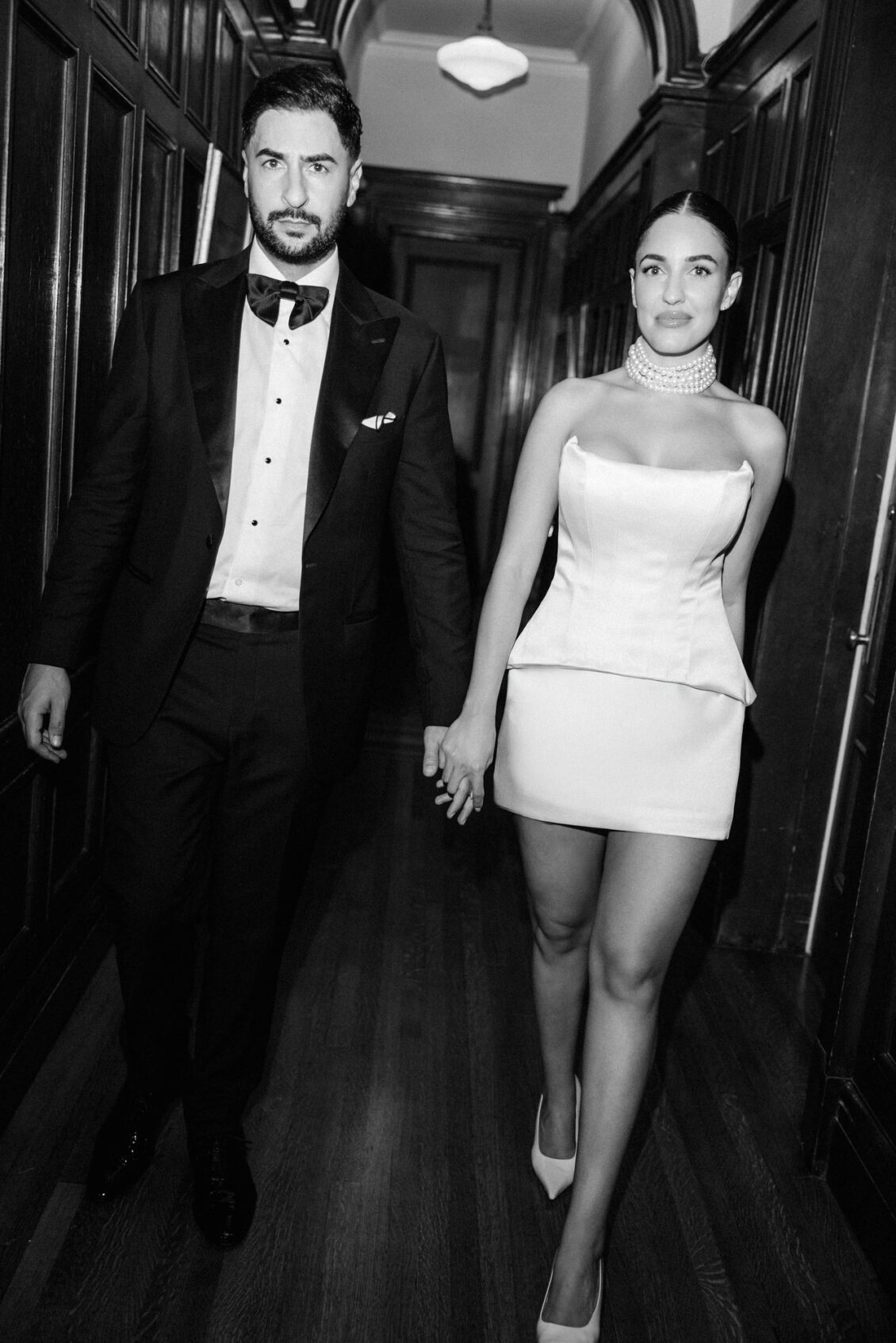 Black and white photo of Lara in a white mini dress and pearls walking hand in hand with Alex down a wood-panelled hallway