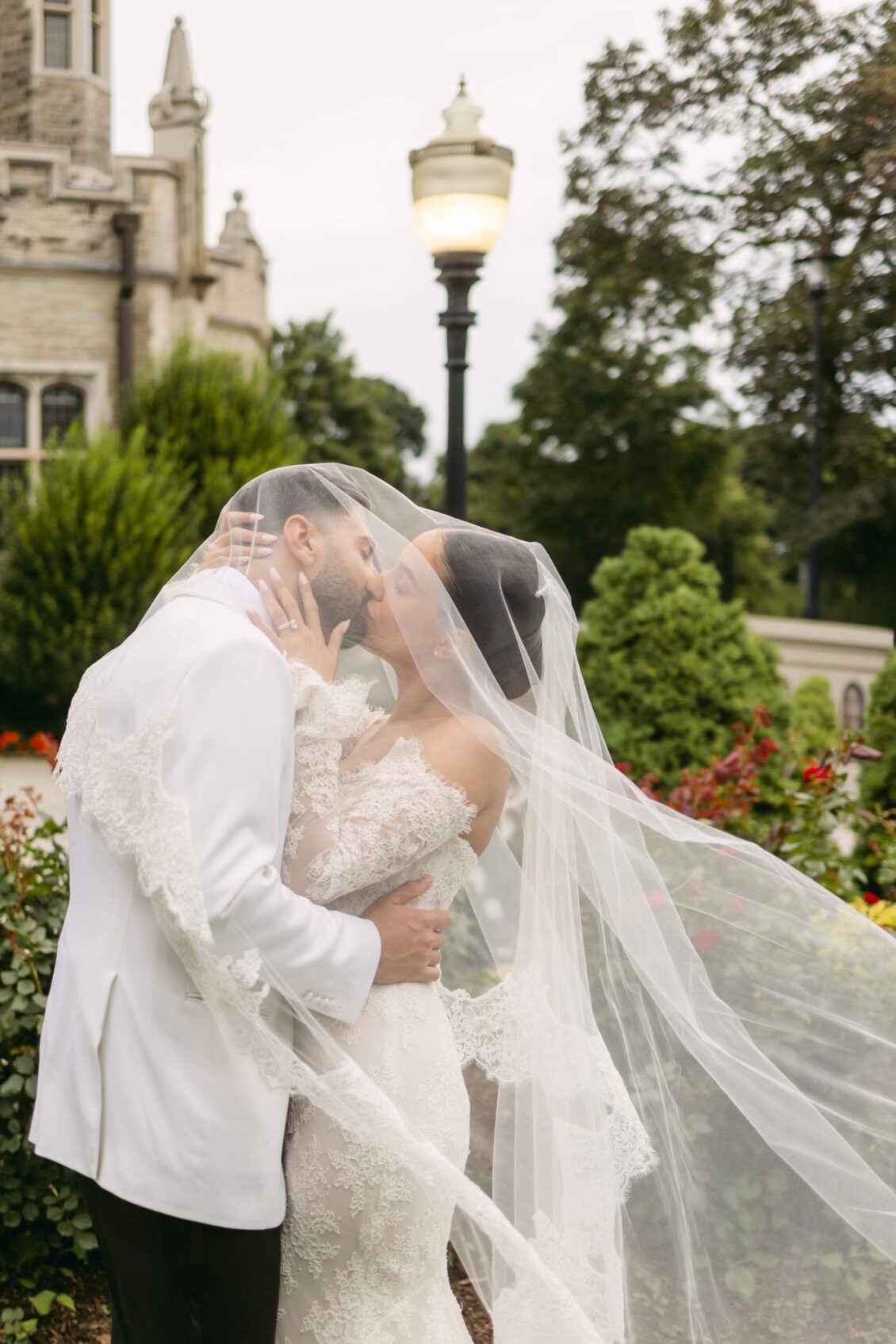 Lara and Alex kiss as her cathedral veil blows around them, with a Casa Loma tower and a lamp post behind
