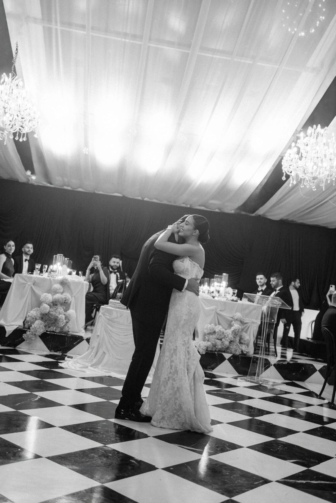 Lara and Alex dance on a checkered marble dance floor under crystal chandeliers as guests watch