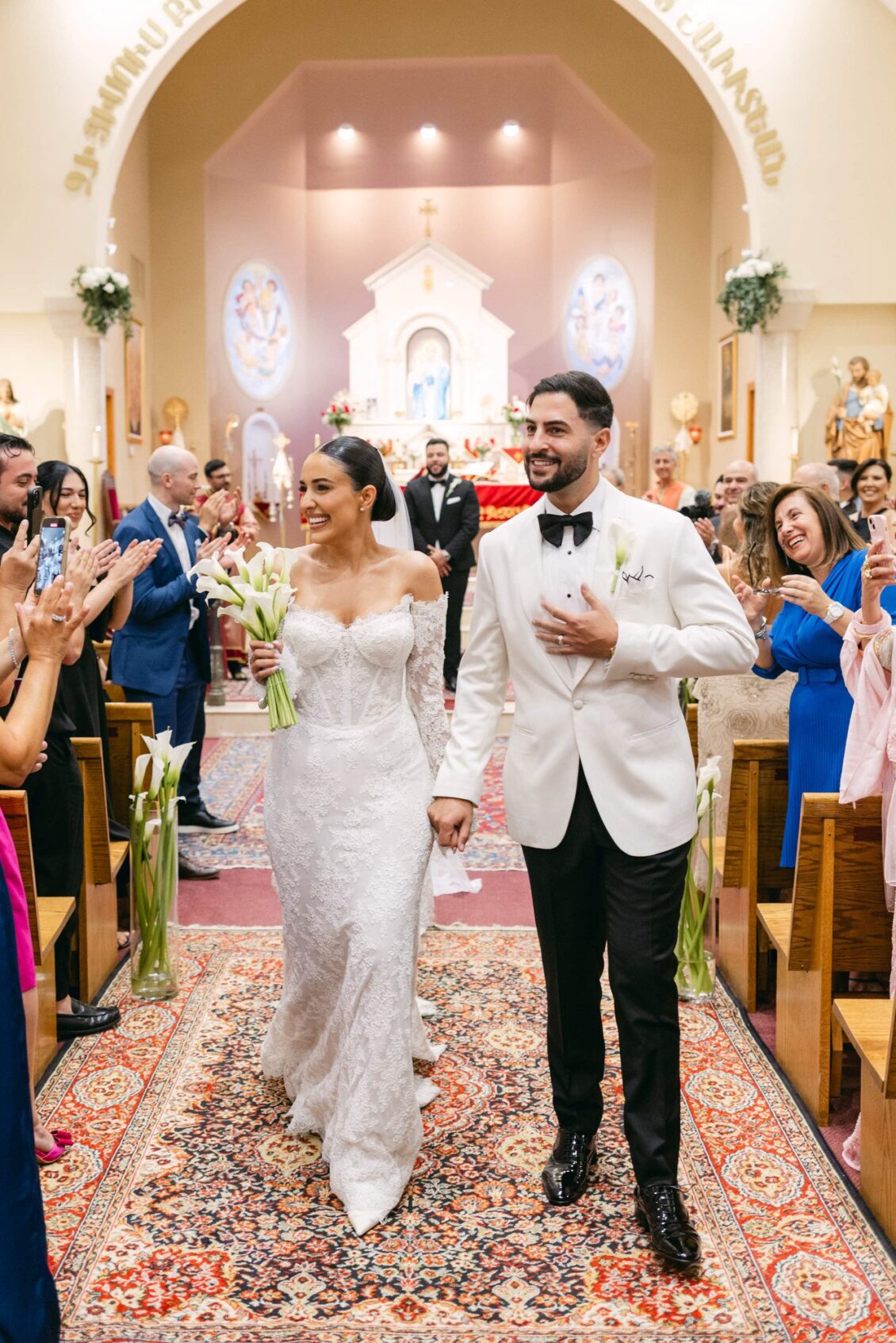 Lara and Alex walk back up the aisle on a patterned carpet as guests applaud