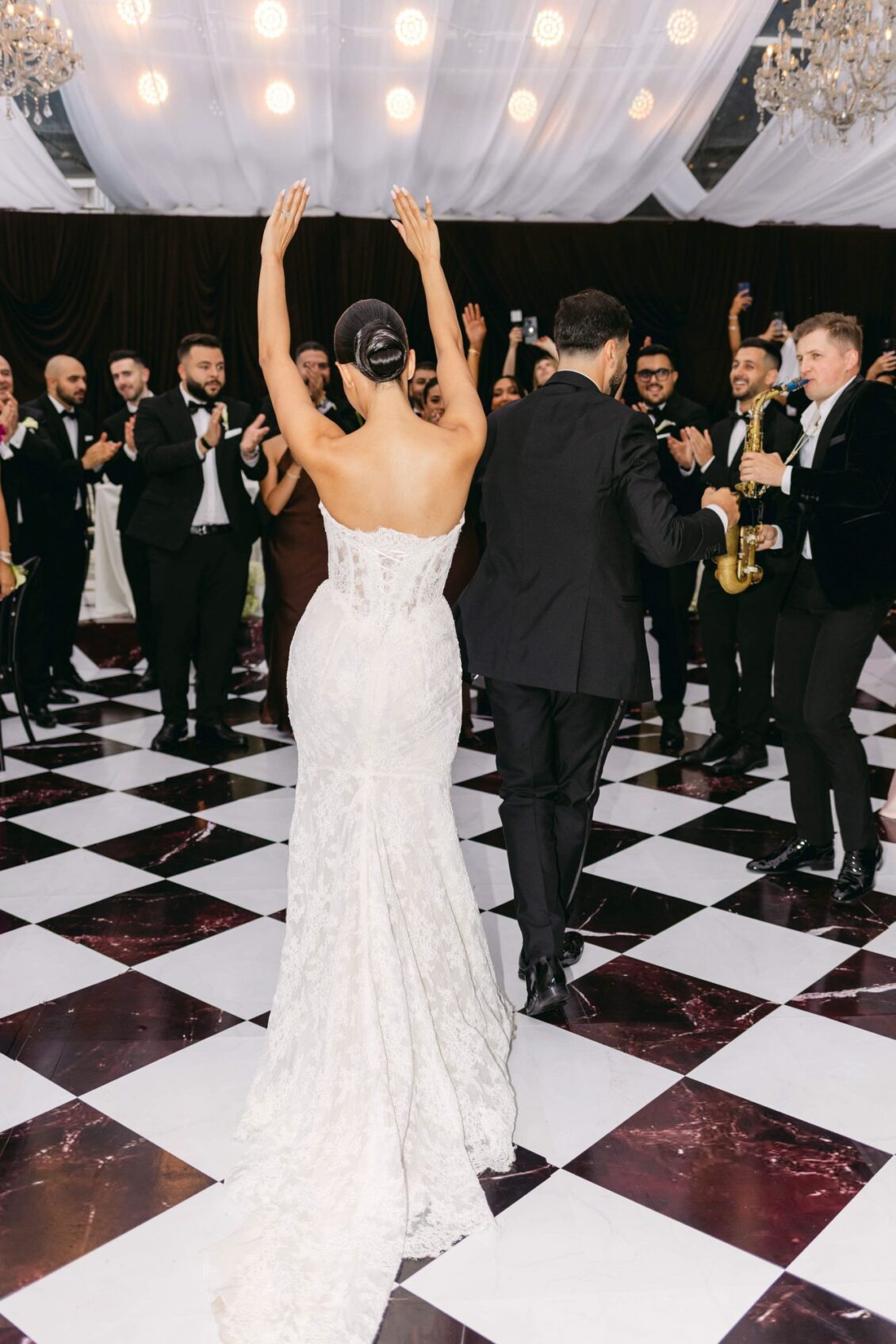 Lara raises her arms as she and Alex step onto the checkered dance floor beside a saxophonist and cheering guests