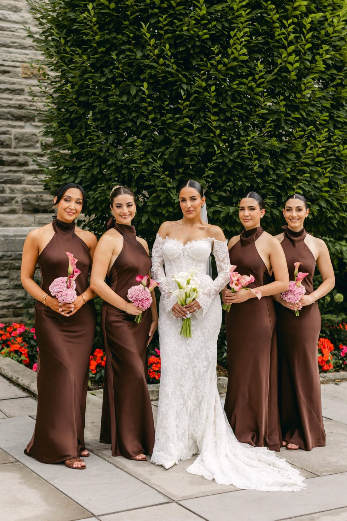 Lara with her bridesmaids in brown satin halter gowns holding pink bouquets in front of a hedge