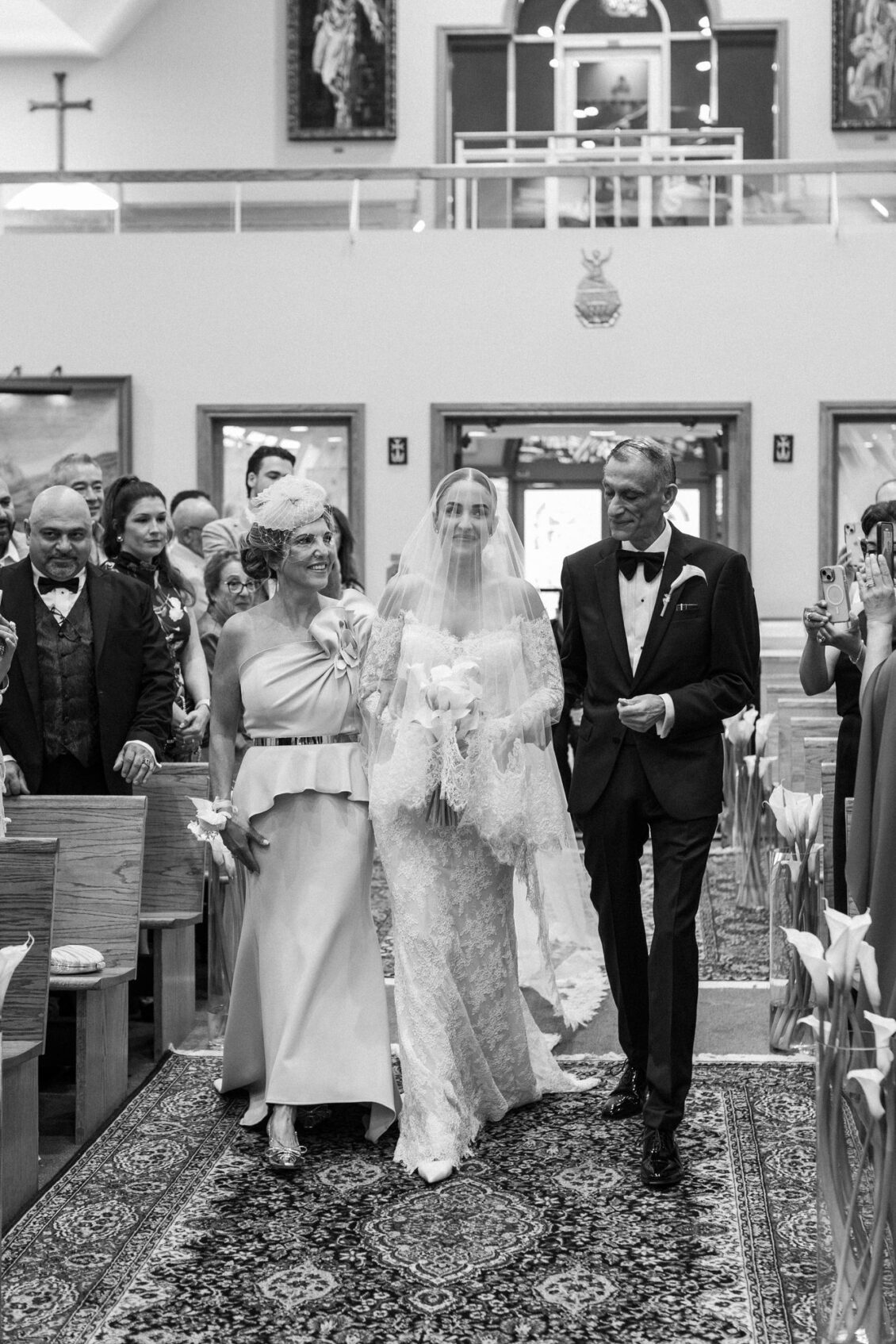 Black and white photo of Lara holding calla lilies as she is escorted down the aisle, her father on one side and a woman in a satin dress on the other