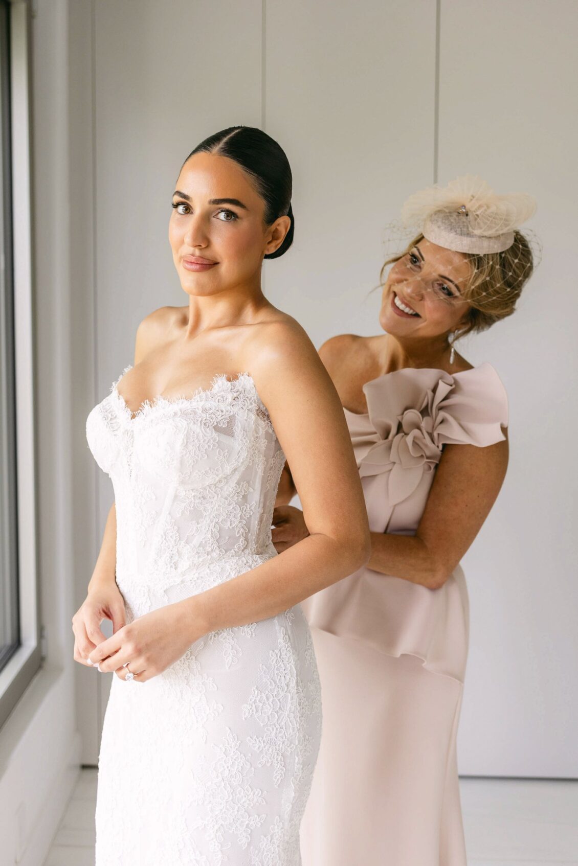 Lara smiles as a woman in a blush dress fastens the back of her strapless lace gown