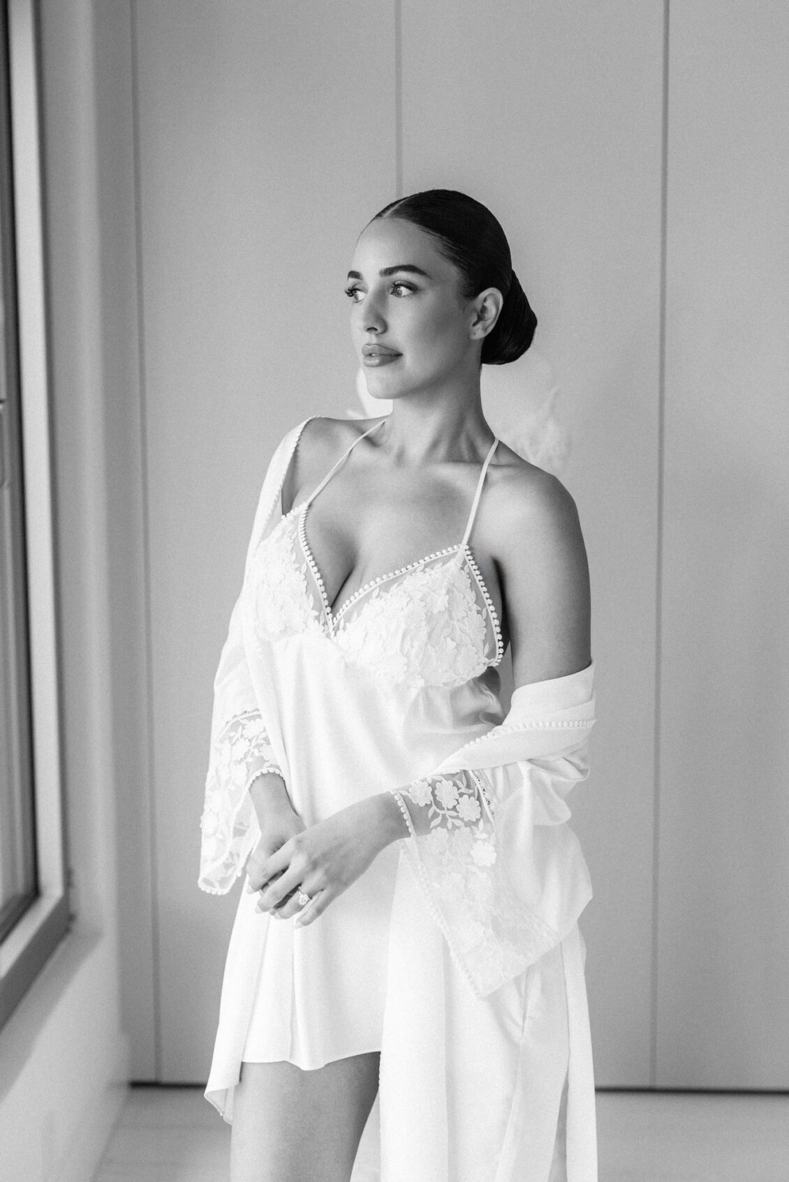 Black and white photo of Lara standing by a tall window in an ivory robe and lace-trimmed chemise