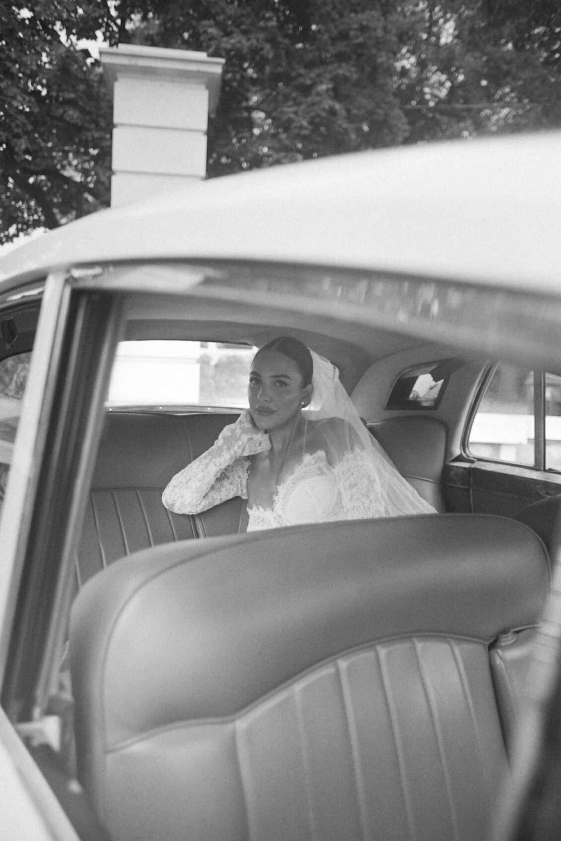 Black and white photo of Lara in her veil seated in the back of a vintage car, seen through the open door