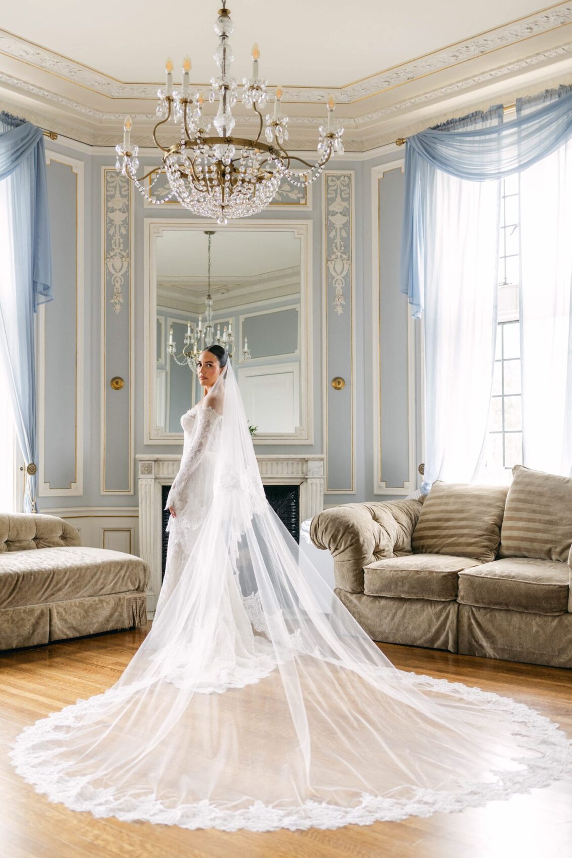 Lara stands in a room with a crystal chandelier and blue drapes, her veil pooled across the parquet floor