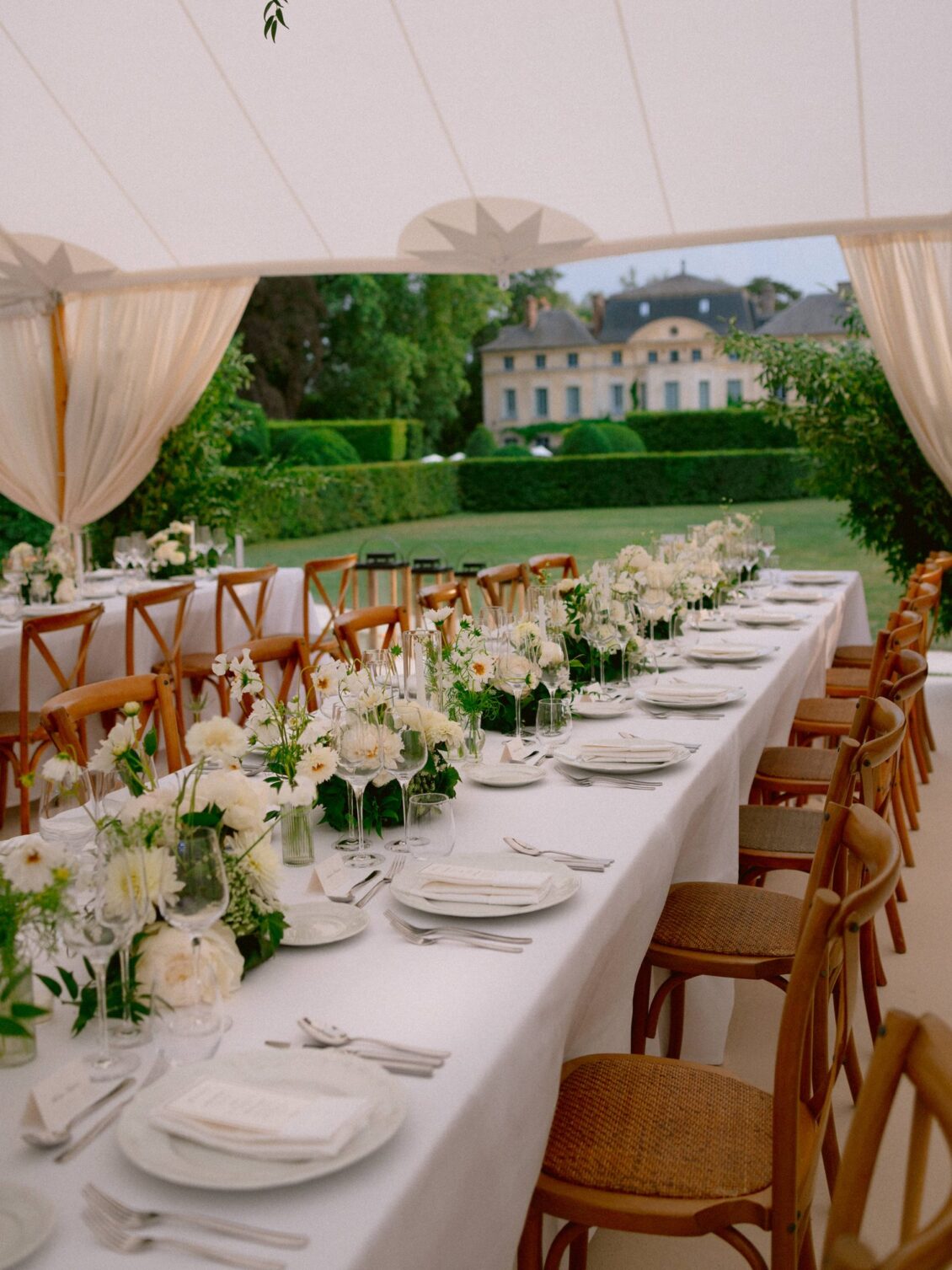 Long white tables with low white florals in the tent, the open drapes framing the château beyond